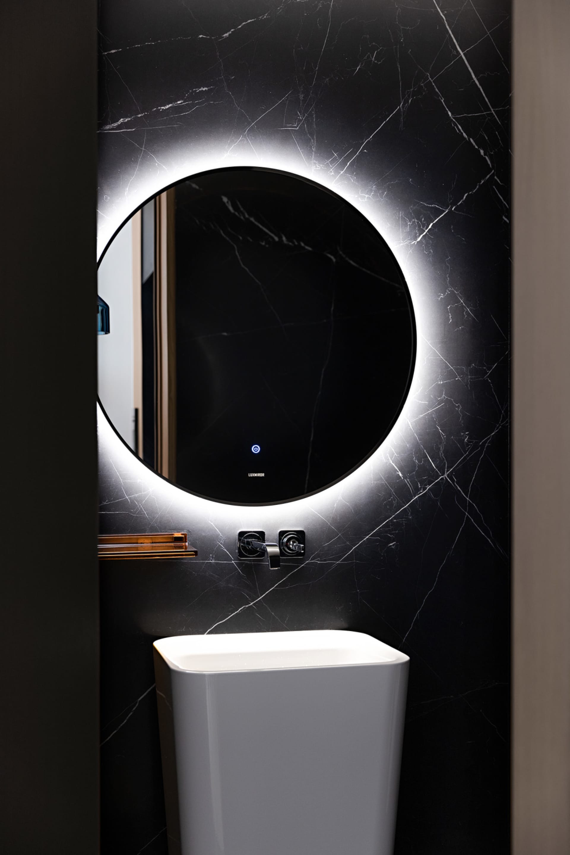 Modern bathroom with a sleek, round mirror with backlit LED lighting and a white ceramic pedestal sink against a black marble