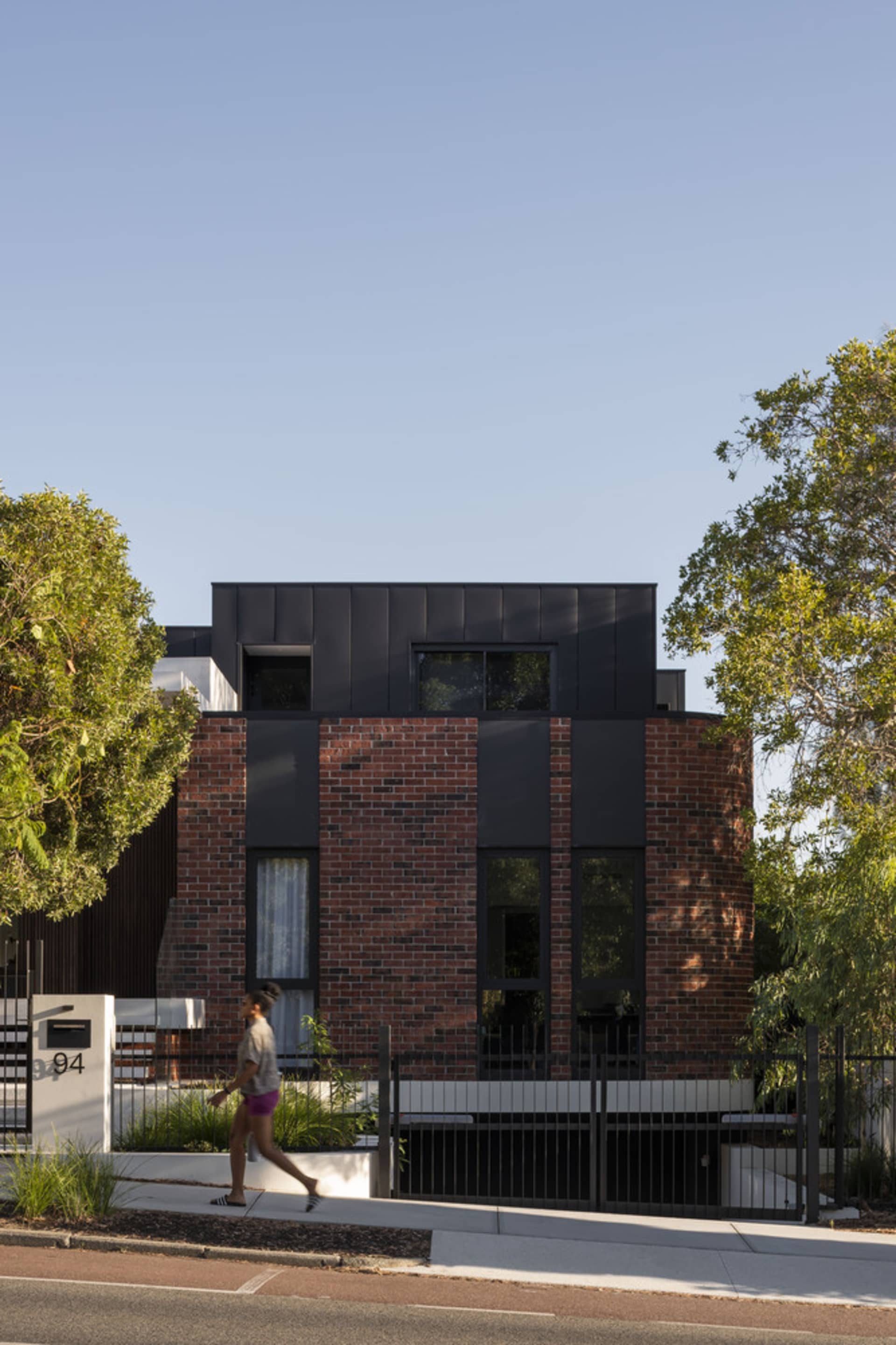 A modern residential house with a brick facade and black accents, set against a clear blue sky with lush green trees surround