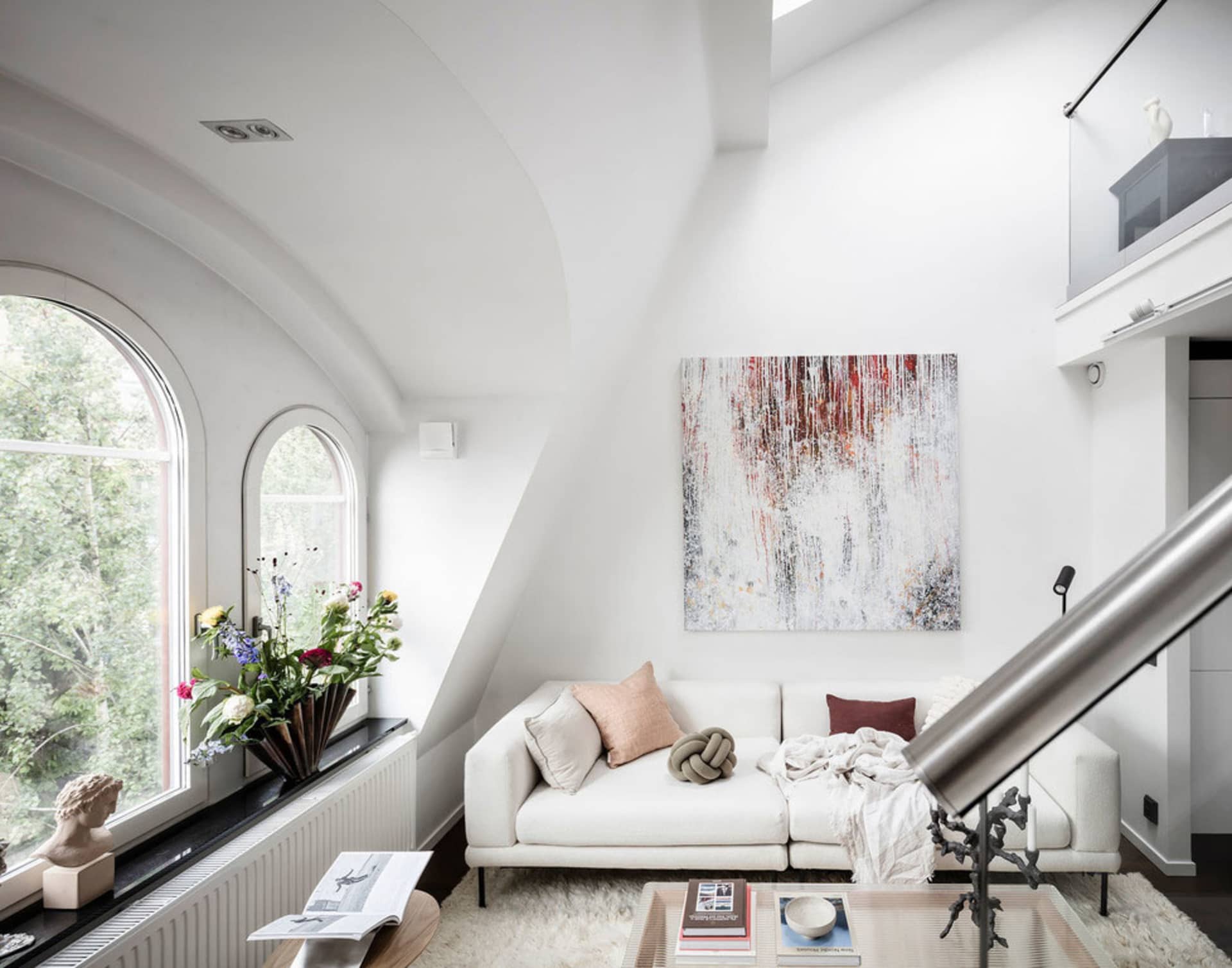 Modern minimalist living room with a white sofa, abstract art, and arched windows overlooking greenery.
