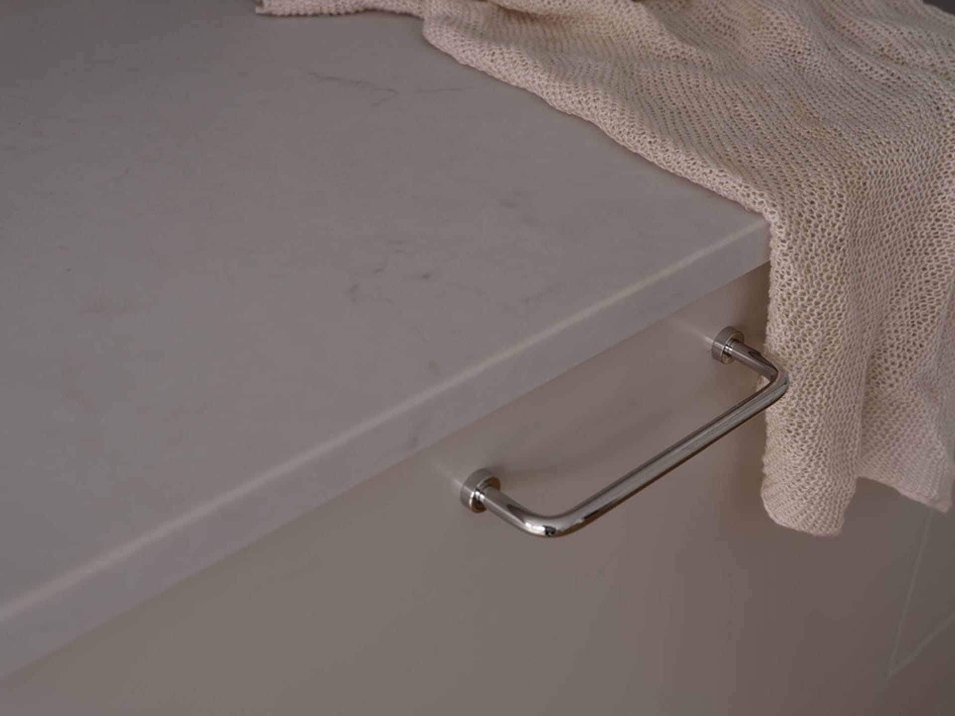 Close-up of a shiny chrome kitchen handle on a marble countertop, with a beige knitted towel draped over the edge.