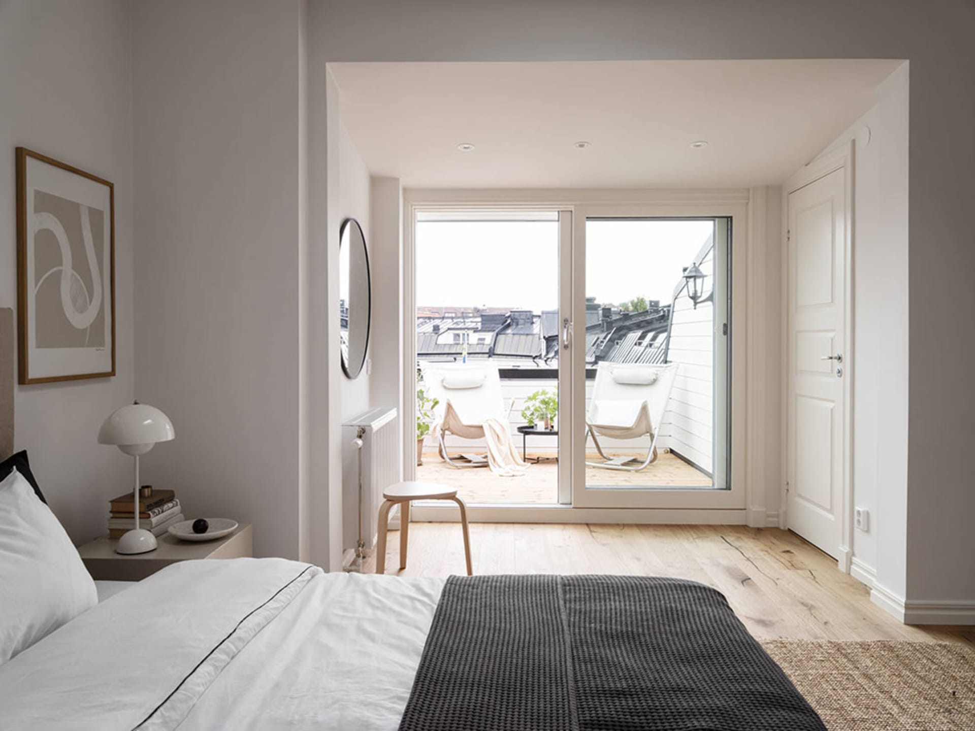 A minimalist bedroom with a wooden floor, white bedding, and a view of a balcony with chairs.