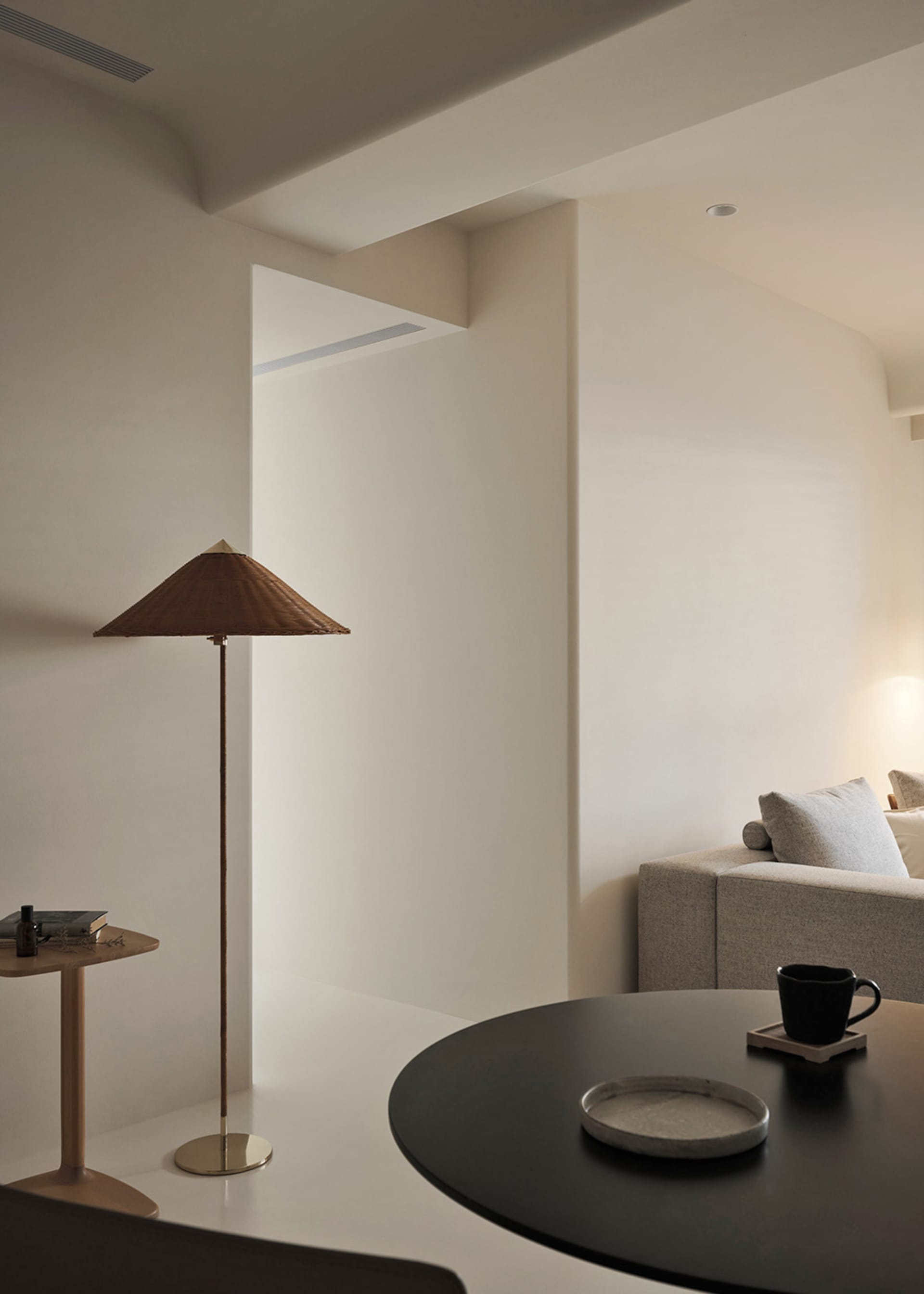 A minimalist living room with a neutral color palette, featuring a sofa, a table with a plate and mug, and a lamp with a coni