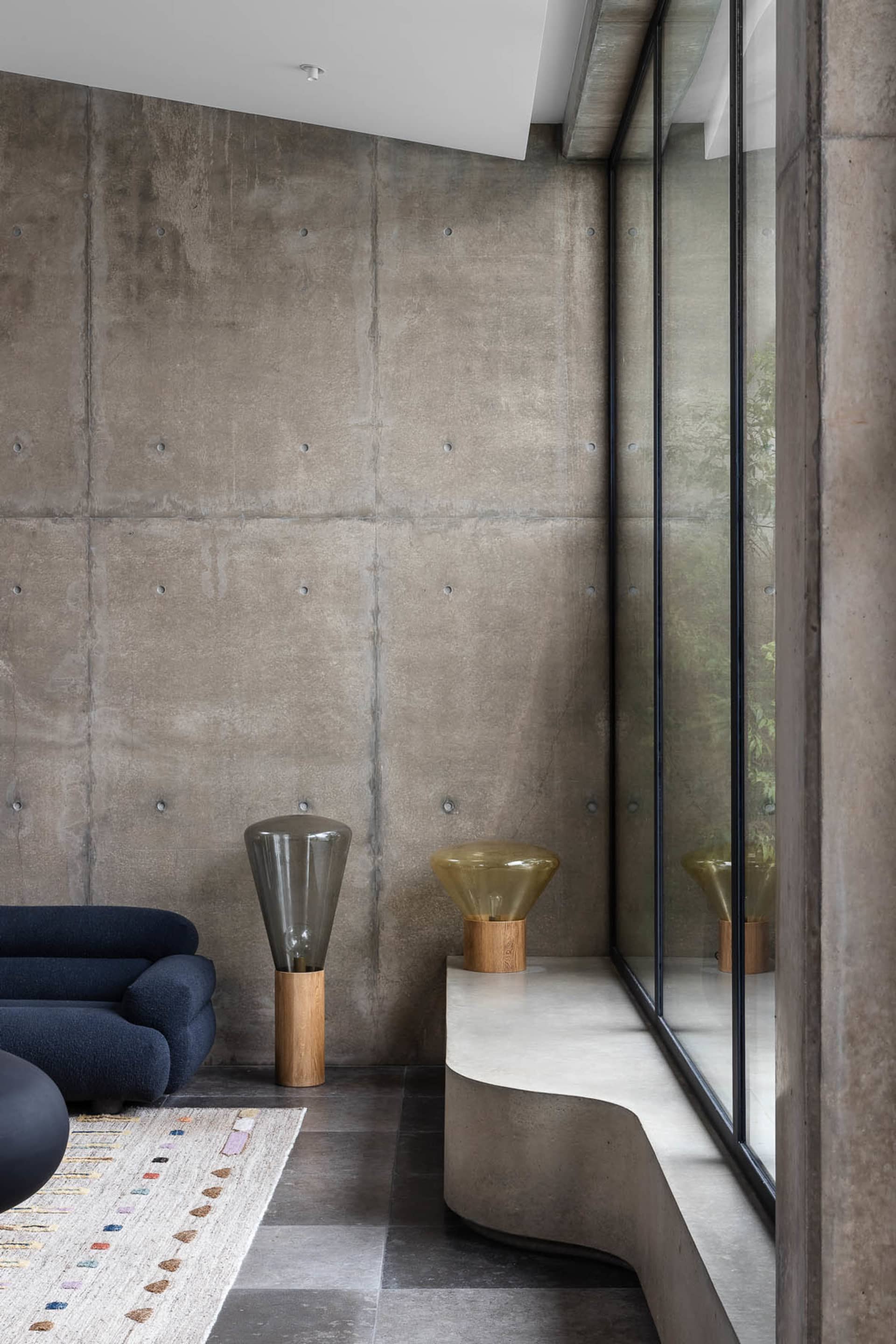 A modern minimalist living room featuring a dark blue sofa, a curved concrete bench, and geometric floor lamp on a patterned