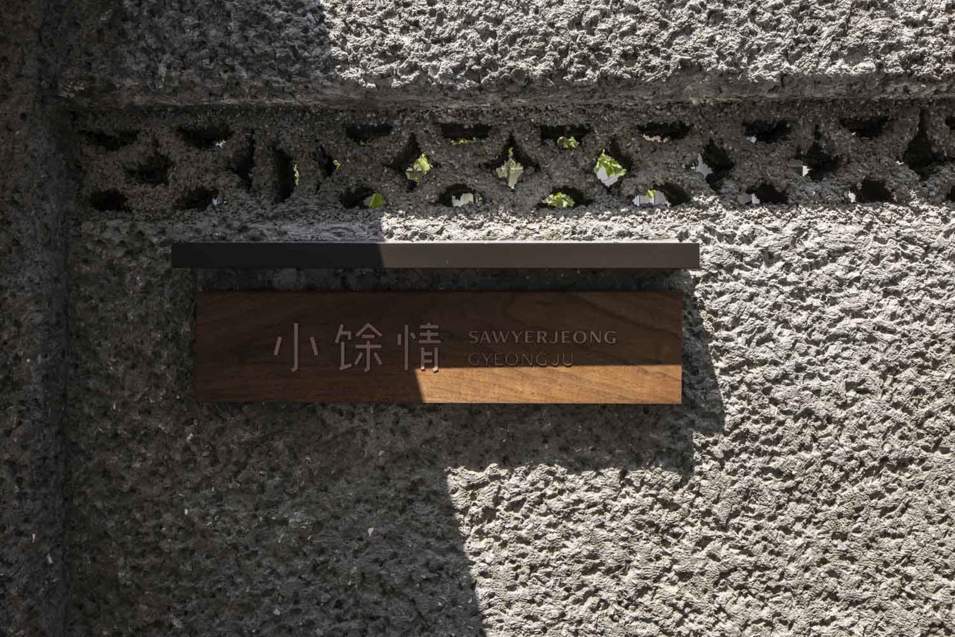 A wooden sign engraved with Chinese characters and English text, mounted on a concrete storefront wall with decorative perfor