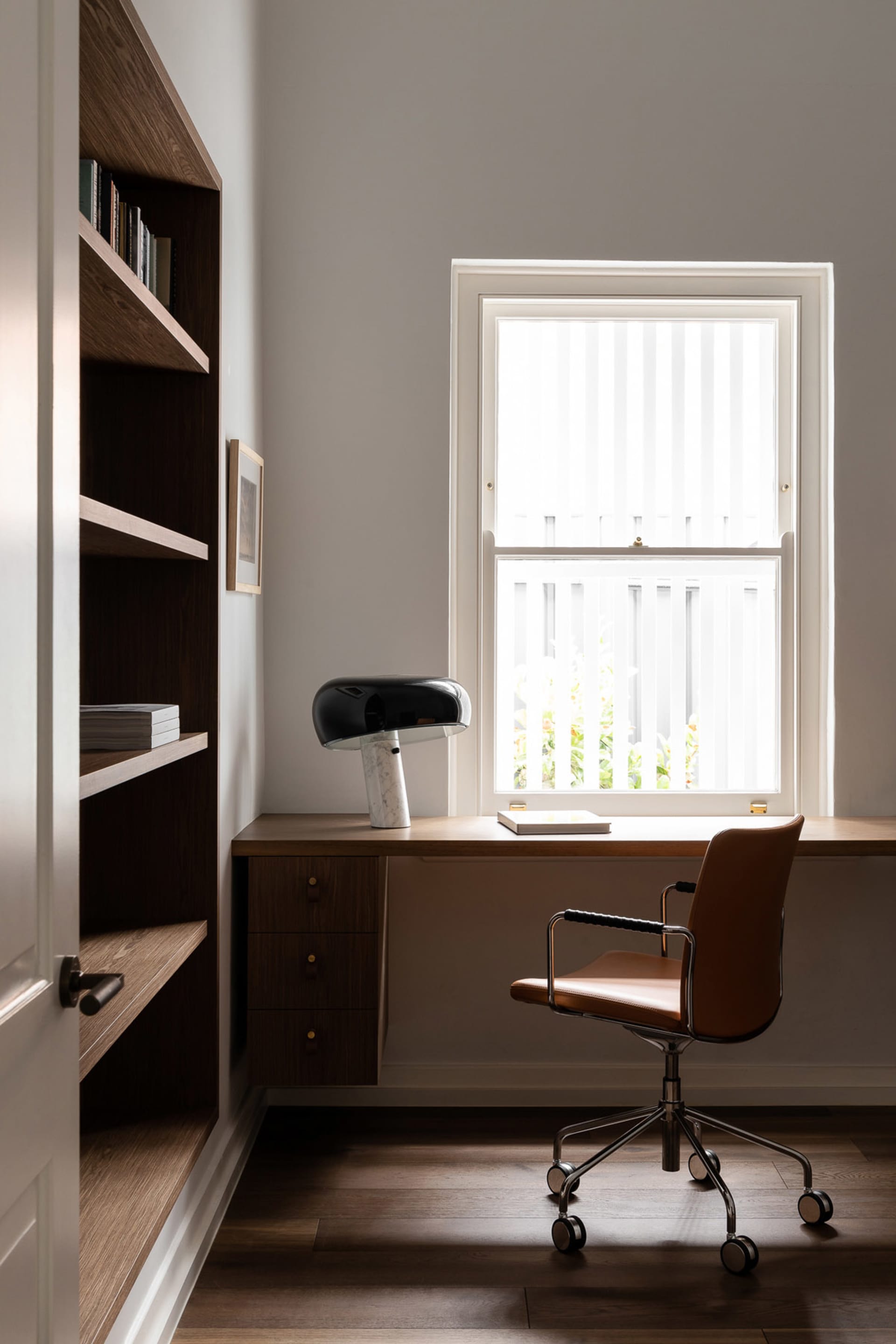 A modern home office with a desk, chair, and lamp next to a bookshelf.