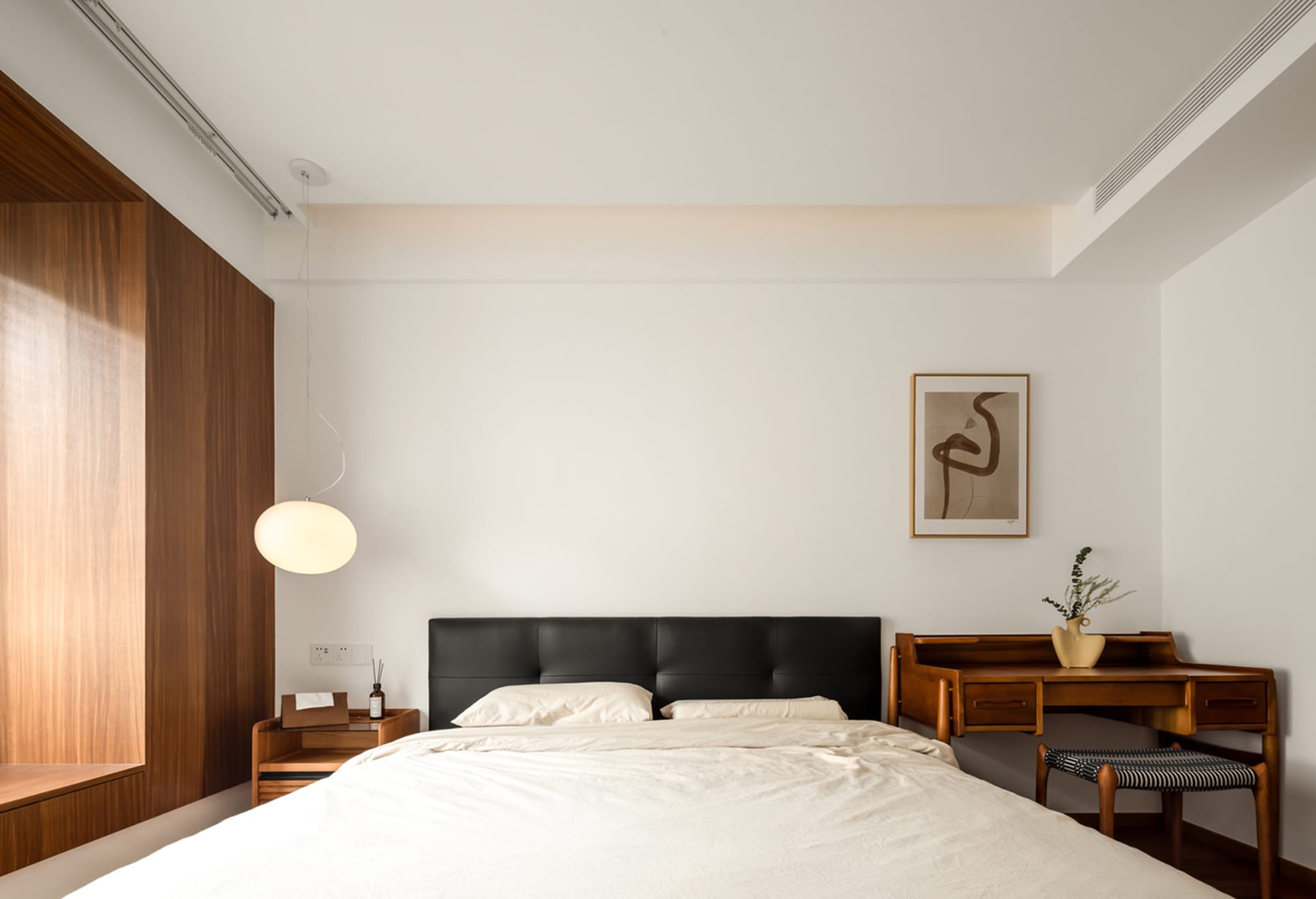 A modern bedroom featuring a black-leather bed with a white comforter and a wooden desk with a plant and vase.