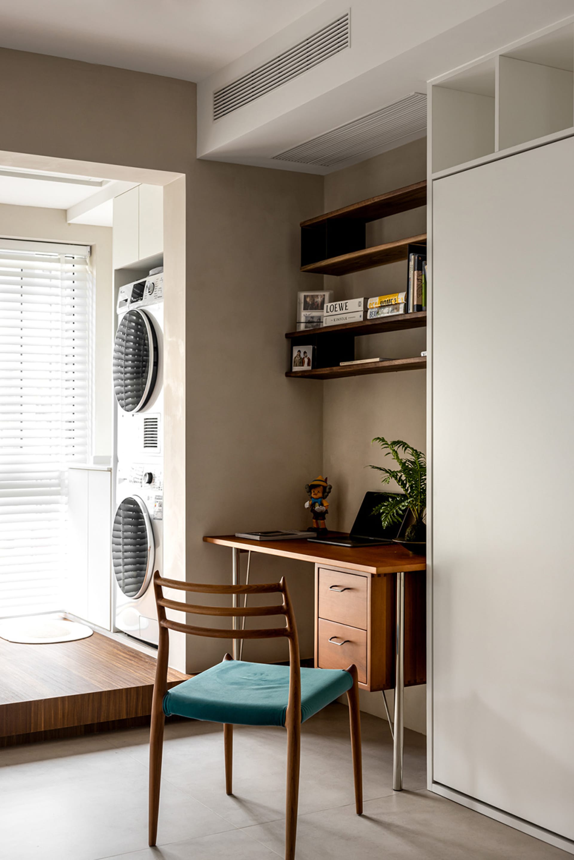 A minimalist home office setup with a wooden desk and chair, featuring a small shelf with books and plants, a clothes wardrob