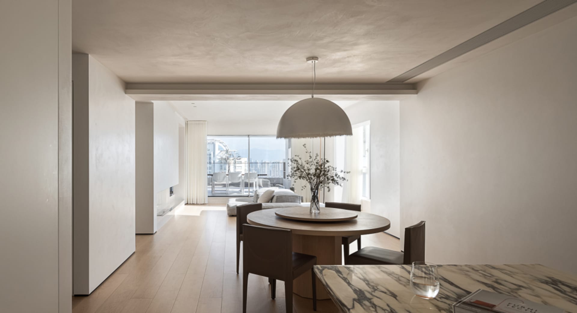 A minimalist dining room with a round wooden table and chairs, accented by a modern hanging light fixture and a vase with flo