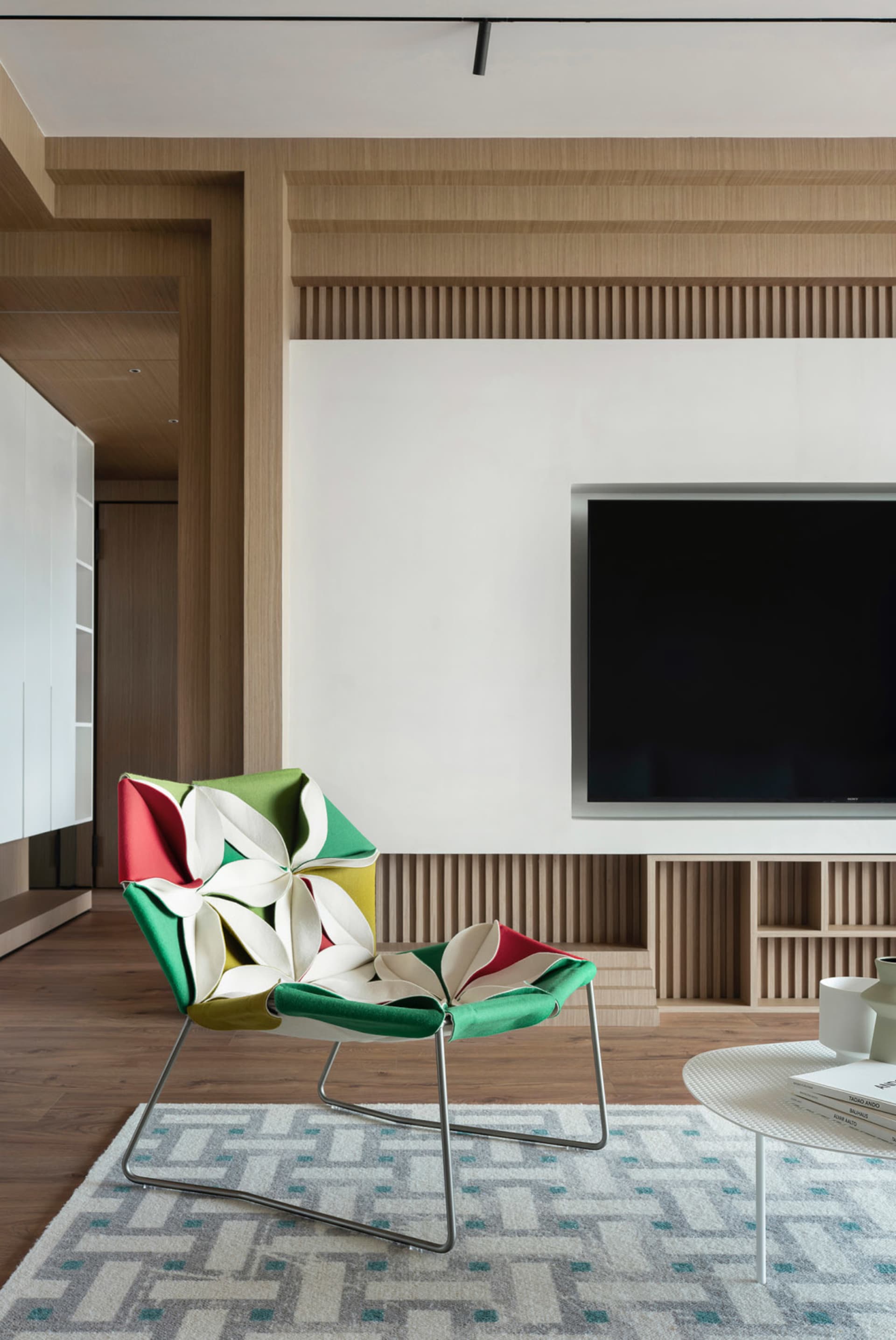 A modern living room featuring a colorful fabric chair with metal legs, a patterned rug, and a large flat-screen TV mounted o