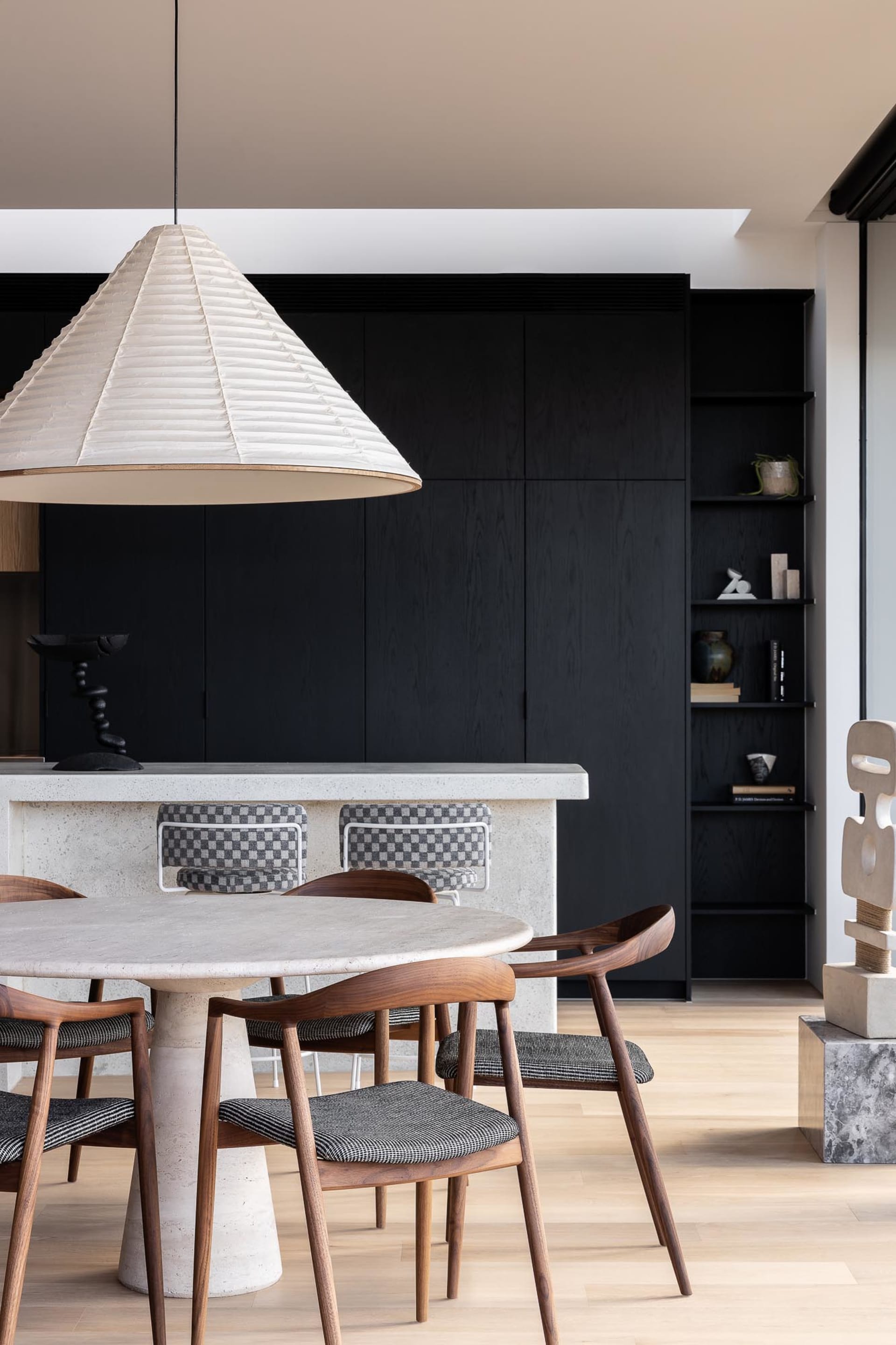 A modern dining room featuring a concrete table and chairs, with a large lampshade overhead and a black shelving unit in the