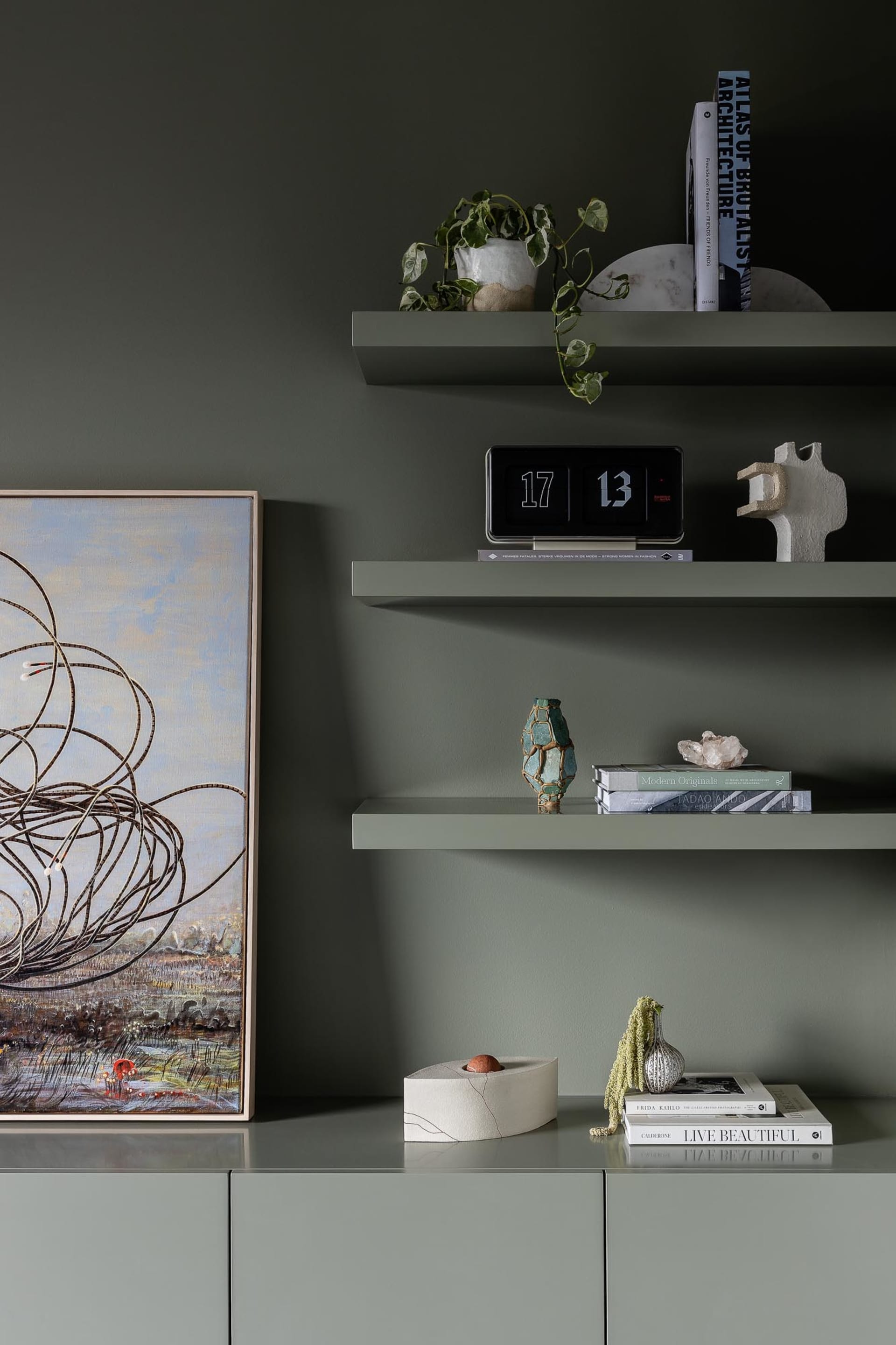 Modern floating shelves with green accents featuring vases, a clock, books, and plants against a dark green wall.