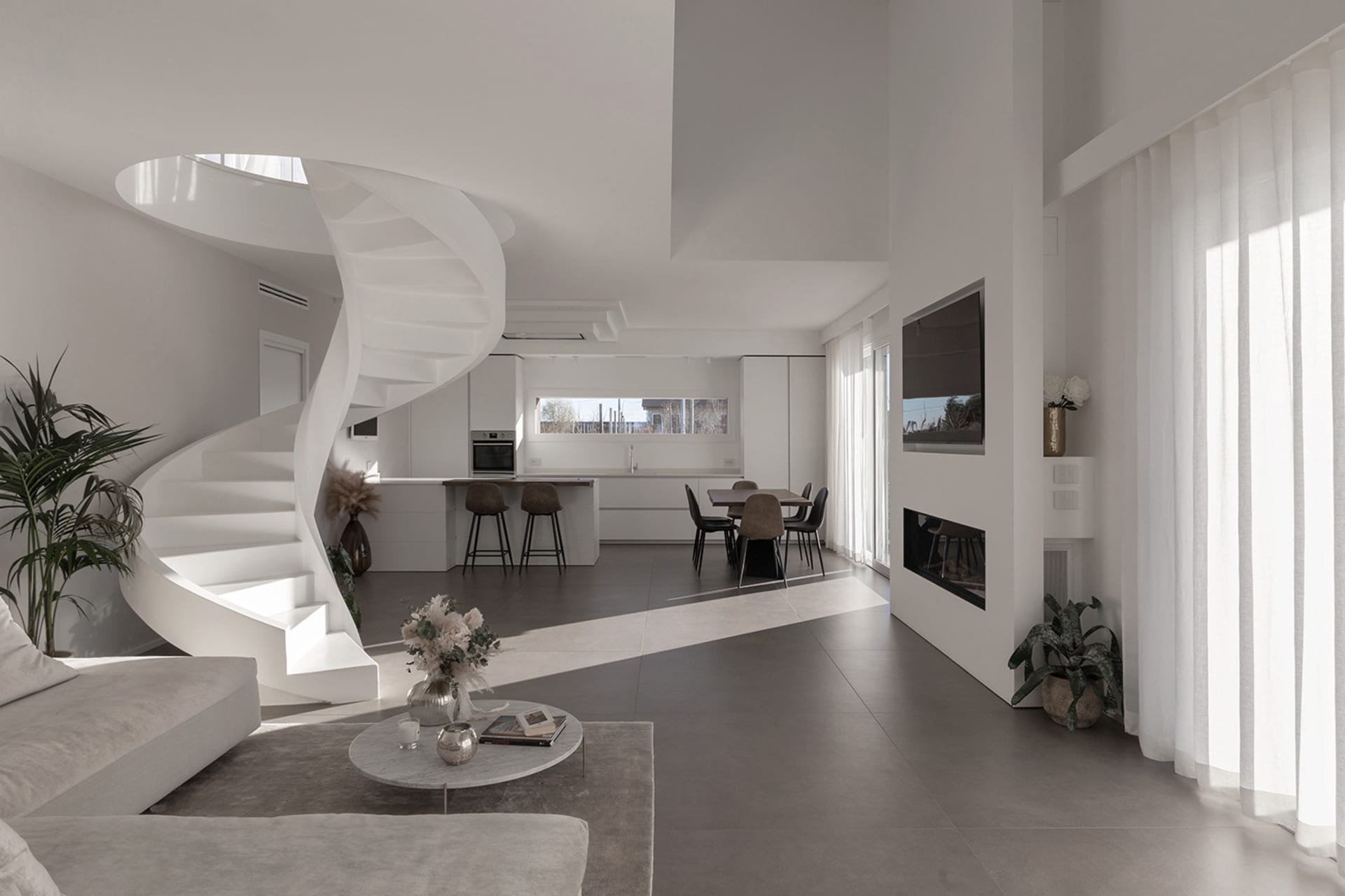 A modern living room featuring a white stone floor, a spiral staircase, a round coffee table, a minimalist sofa, and a kitche