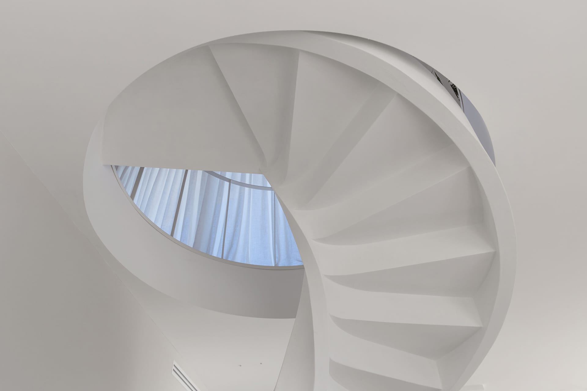 A white, minimalist, concrete staircase with a circular window in the ceiling.