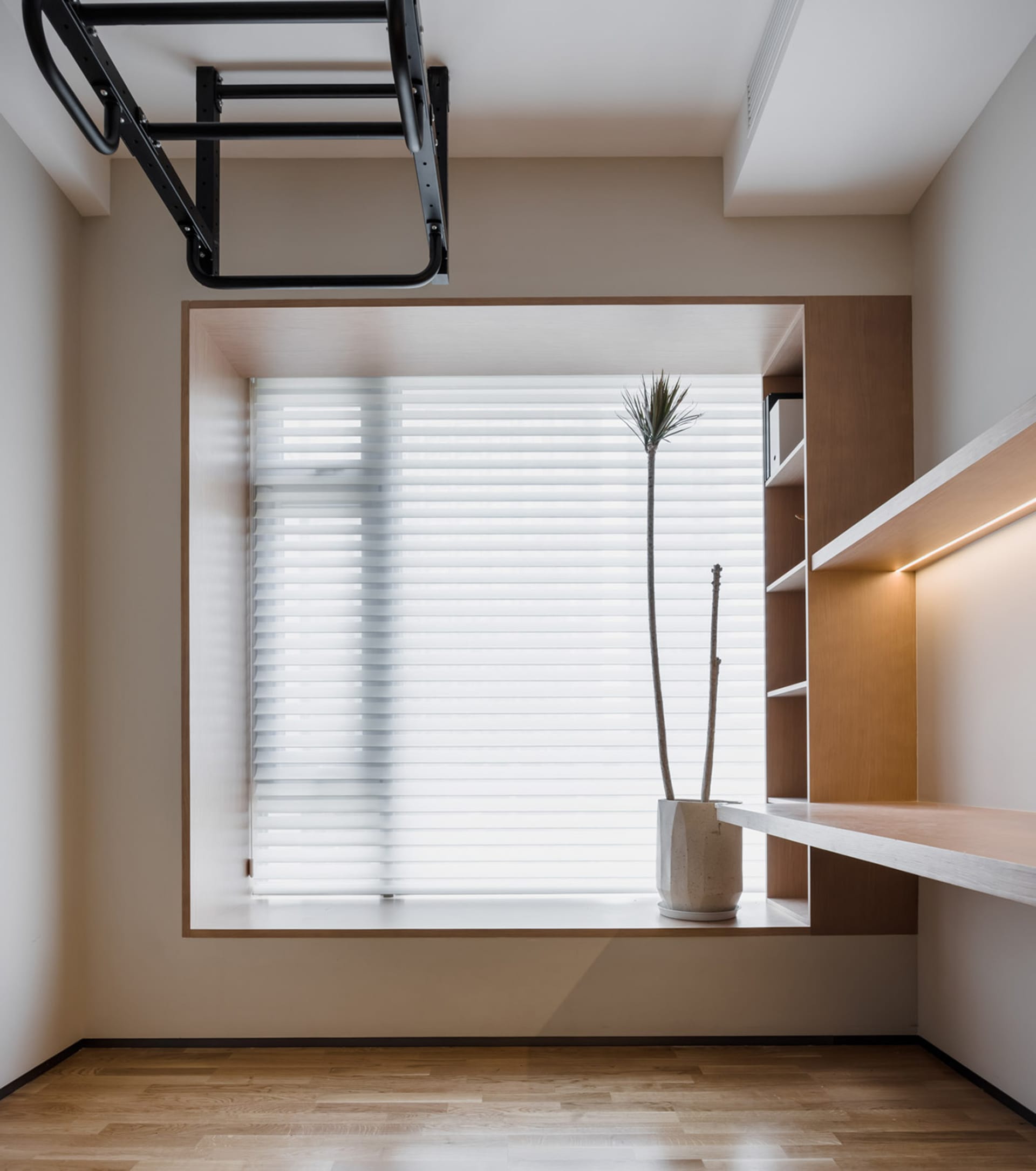 A modern minimalist home office featuring a wooden floor, blinds, potted plants, and a shelf.