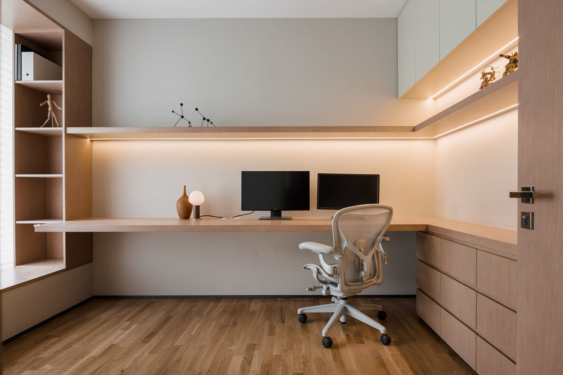 A minimalist office space featuring a wooden desk and chair, with integrated shelving and lighting.
