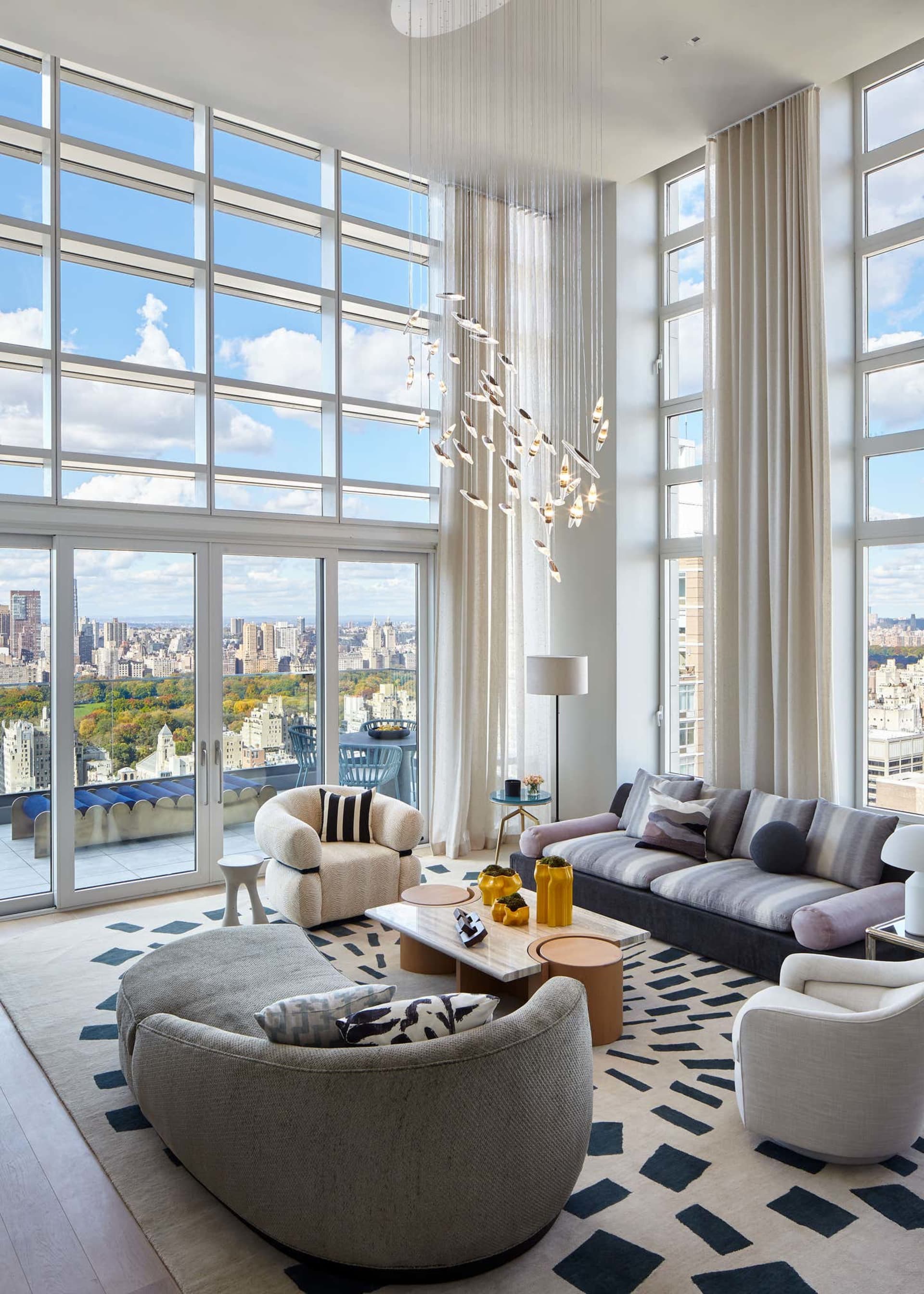 Modern living room with large windows overlooking a cityscape, featuring contemporary furniture and a chandelier.
