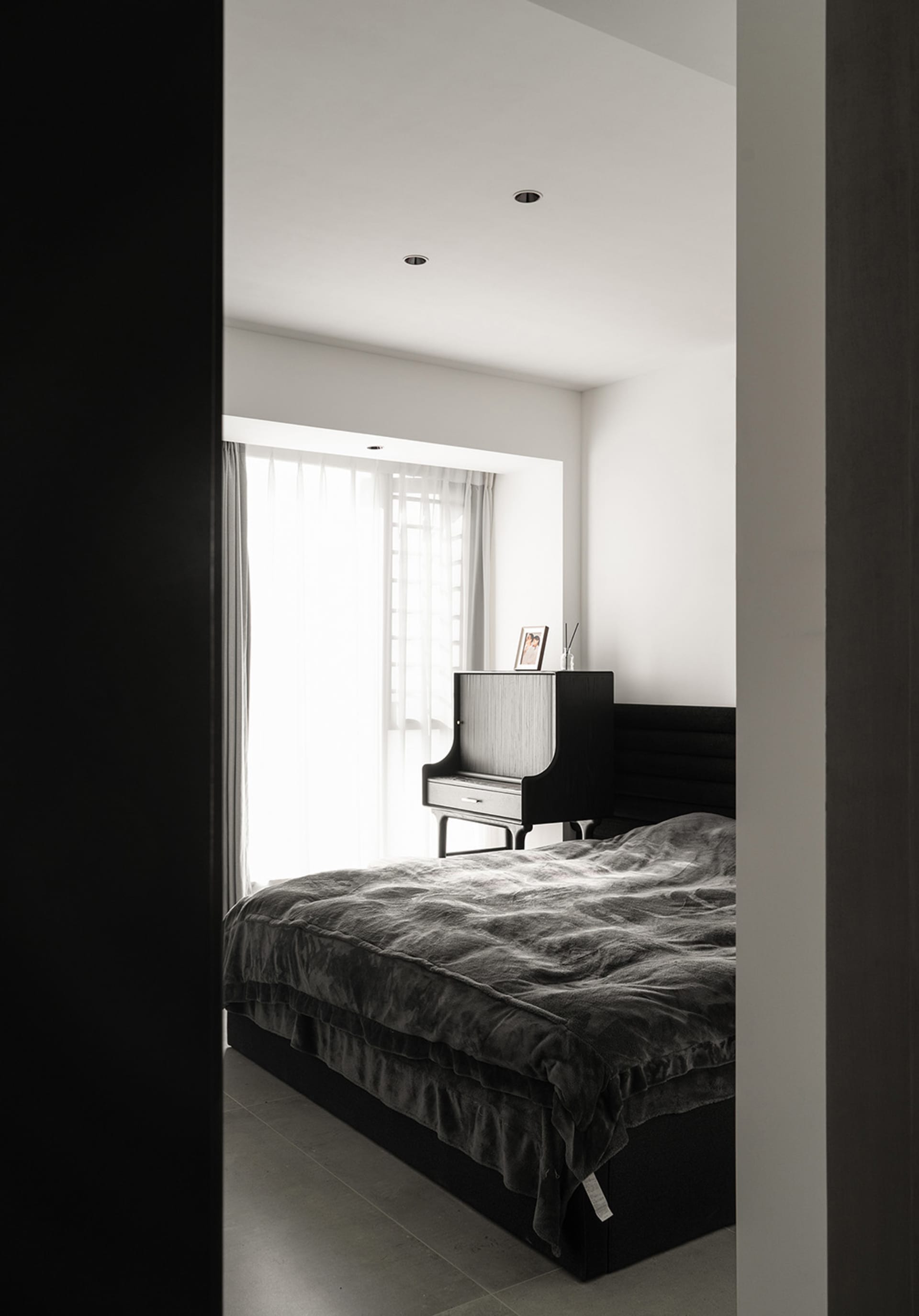 A modern bedroom with a black bed and grey blanket under bright sunlight from a large window.