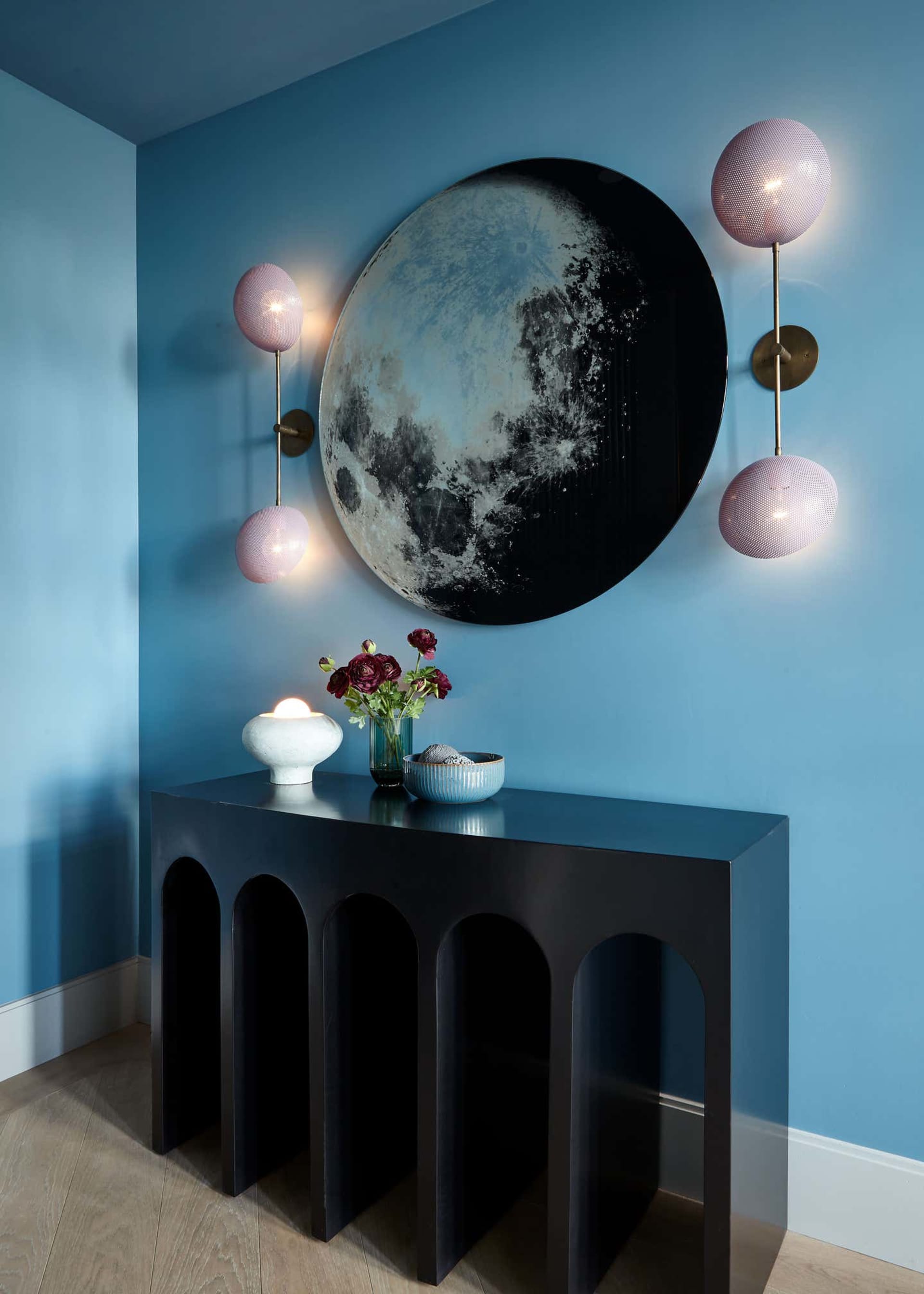 A contemporary living room with blue walls, a black console table with arches, and a wall-mounted art piece of a moon.