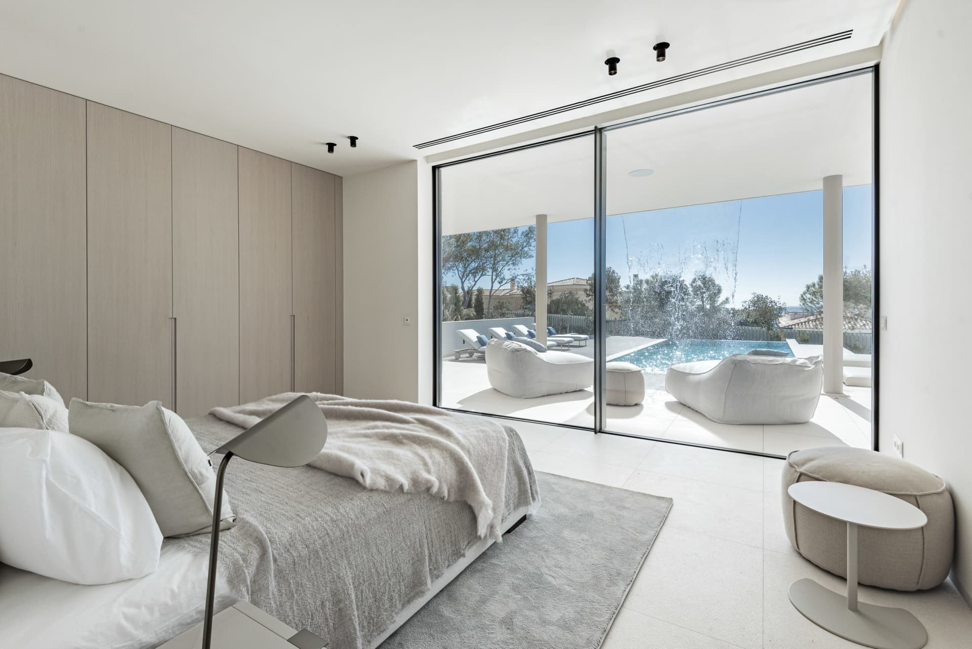 Modern minimalist bedroom with glass walls overlooking a serene outdoor space with a pool and lounge areas.