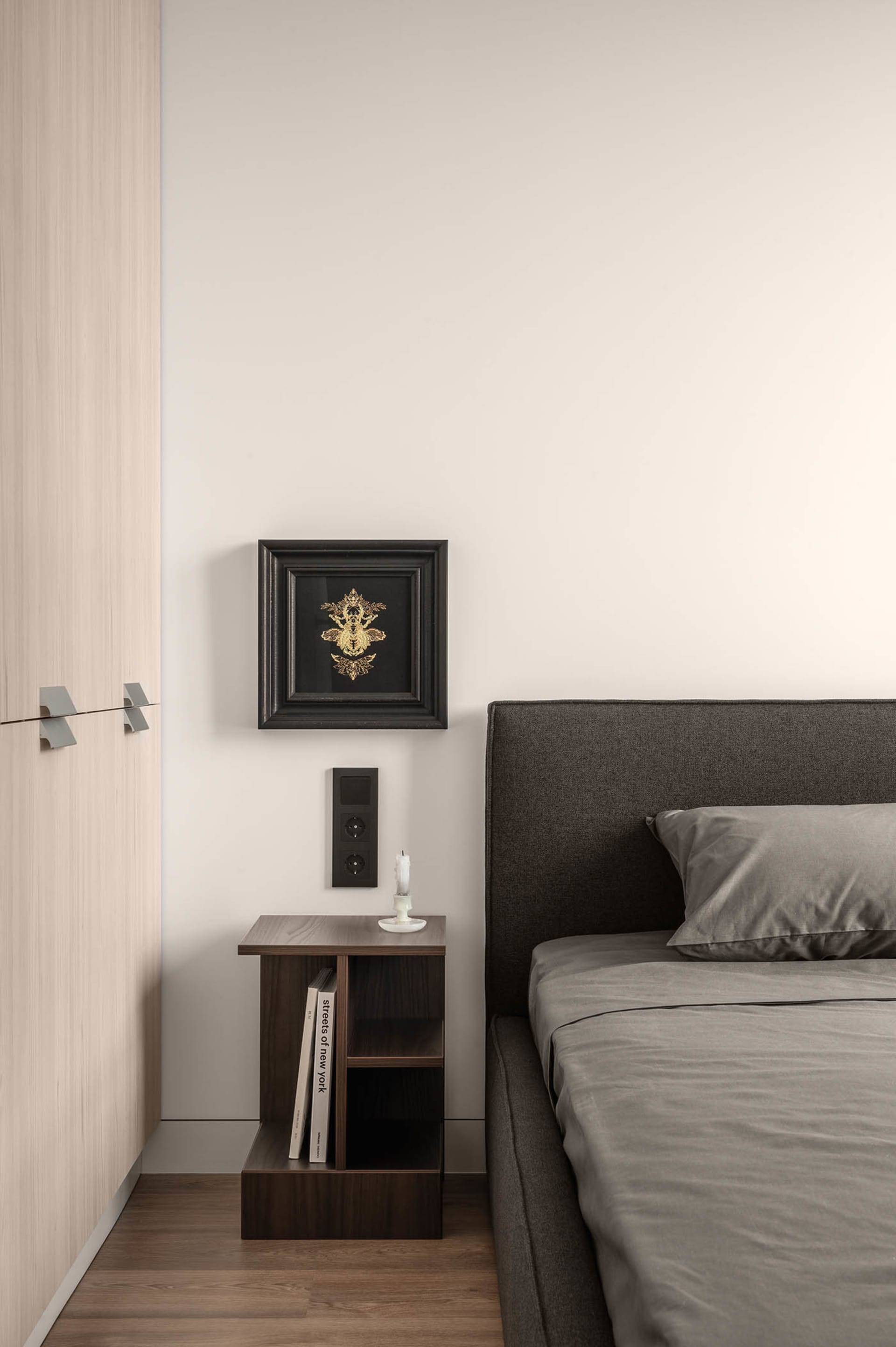 A minimalist bedroom featuring a grey upholstered bed, a wooden bedside table, and a wall-mounted bedside lamp. The decor inc