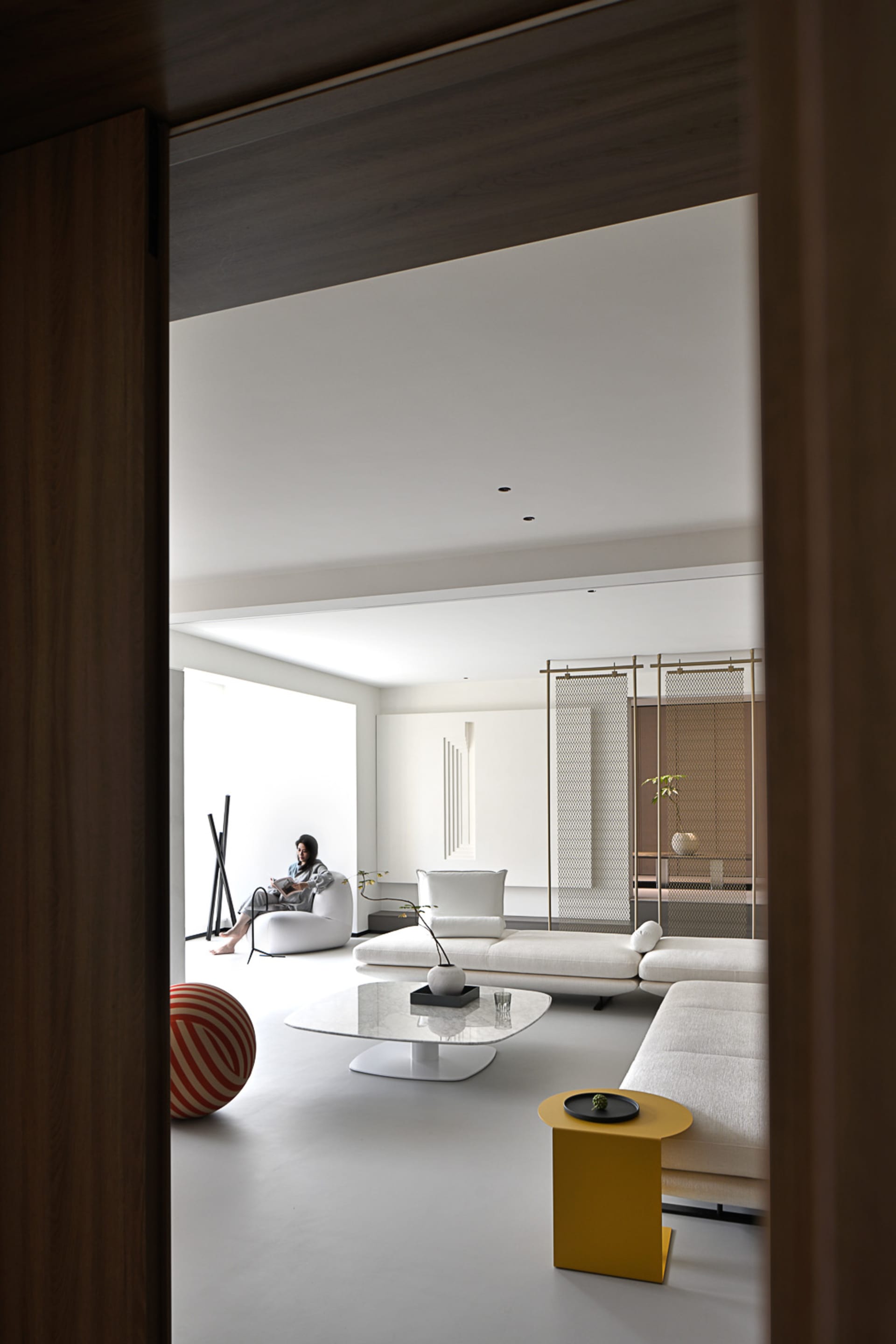 White minimalist living room with lounge sofas, a coffee table, and a round stool, featuring a woman sitting on a white sofa.