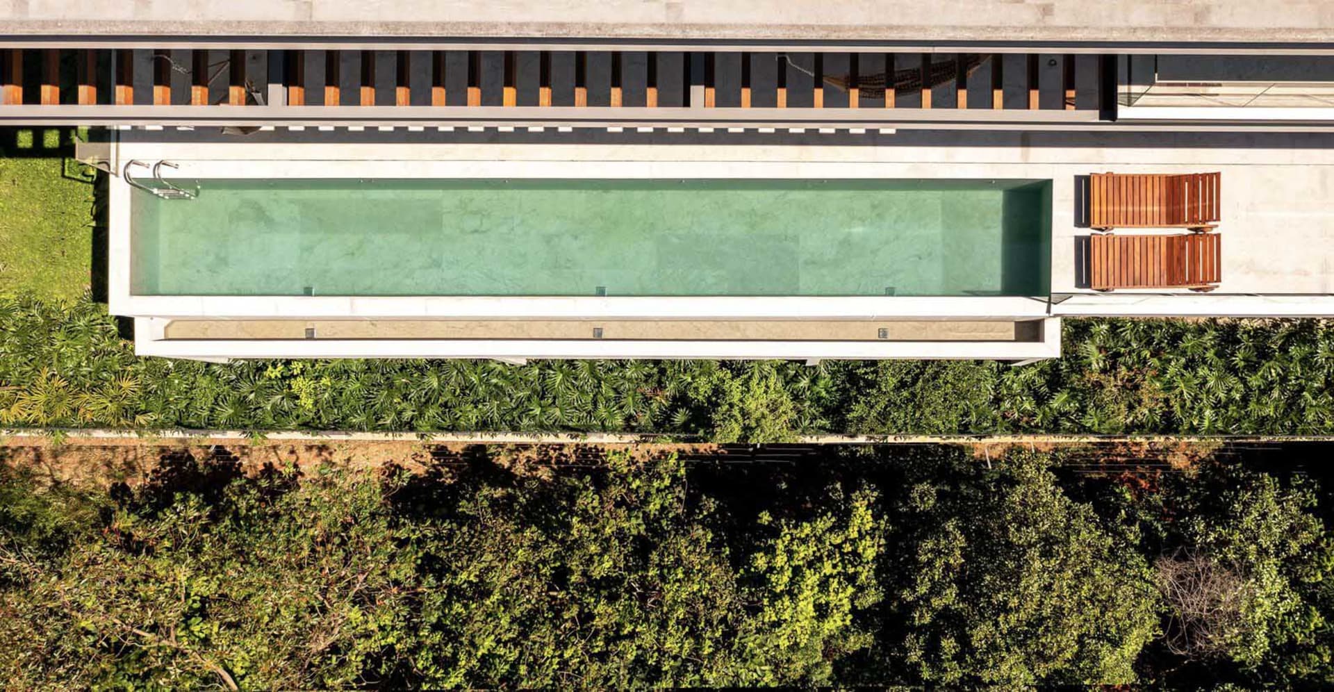 A modern pool with a concrete deck, glass walls, and wooden benches surrounded by greenery.