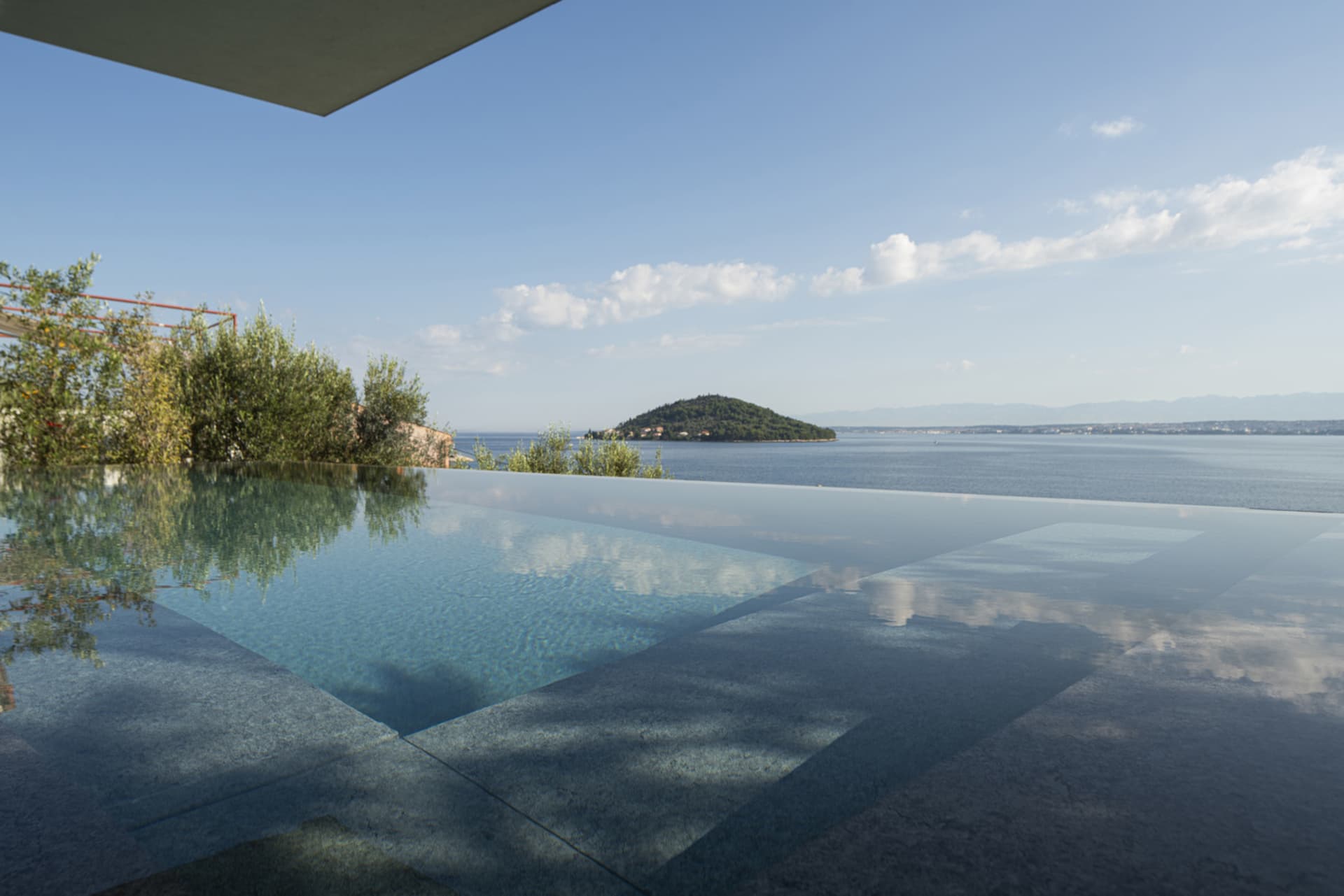 A minimalist swimming pool with a concrete deck and glass walls overlooks a calm sea.