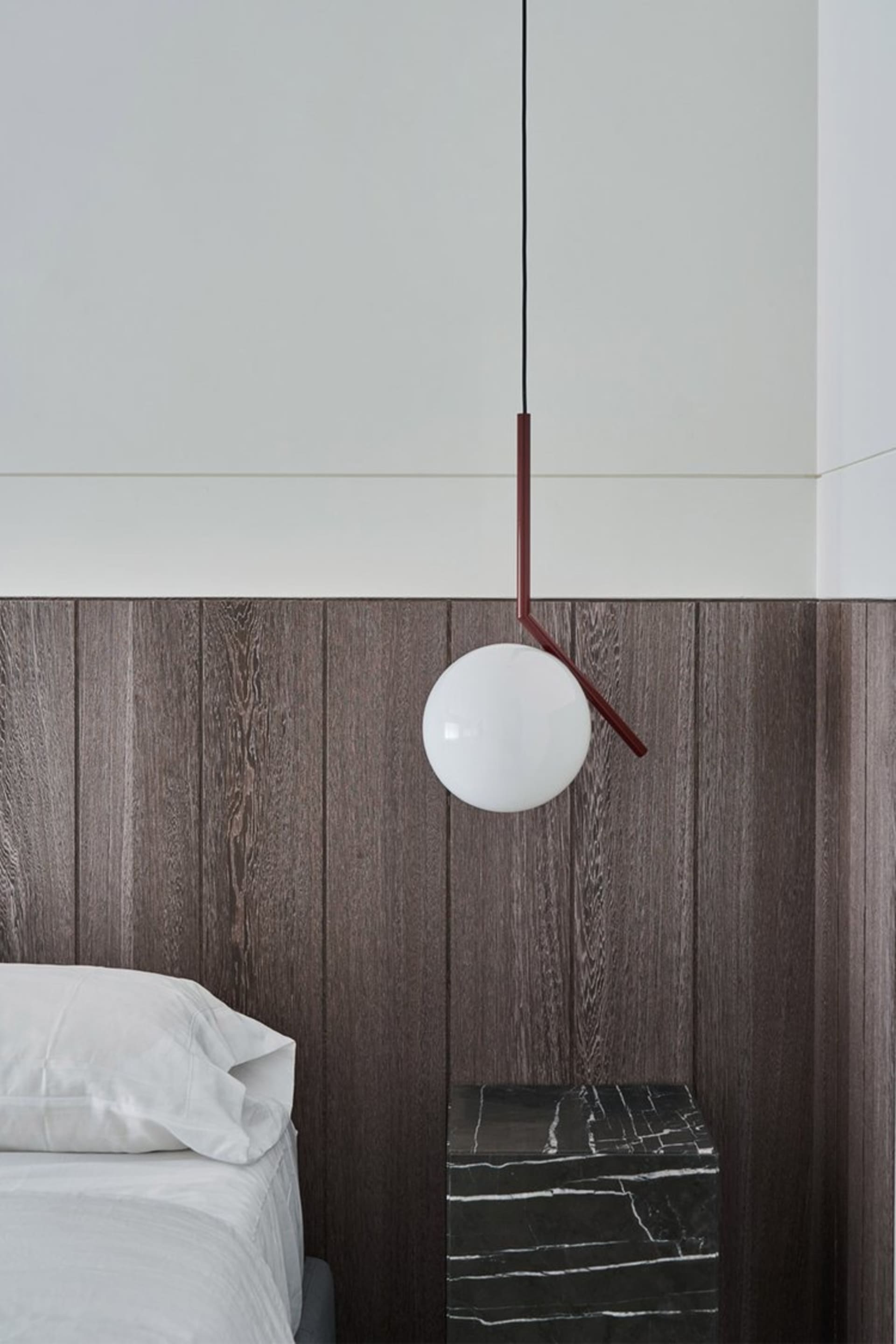 A modern minimalist bedroom featuring a single bed dressed in white bedding, a dark wooden headboard, and a pendant lamp with