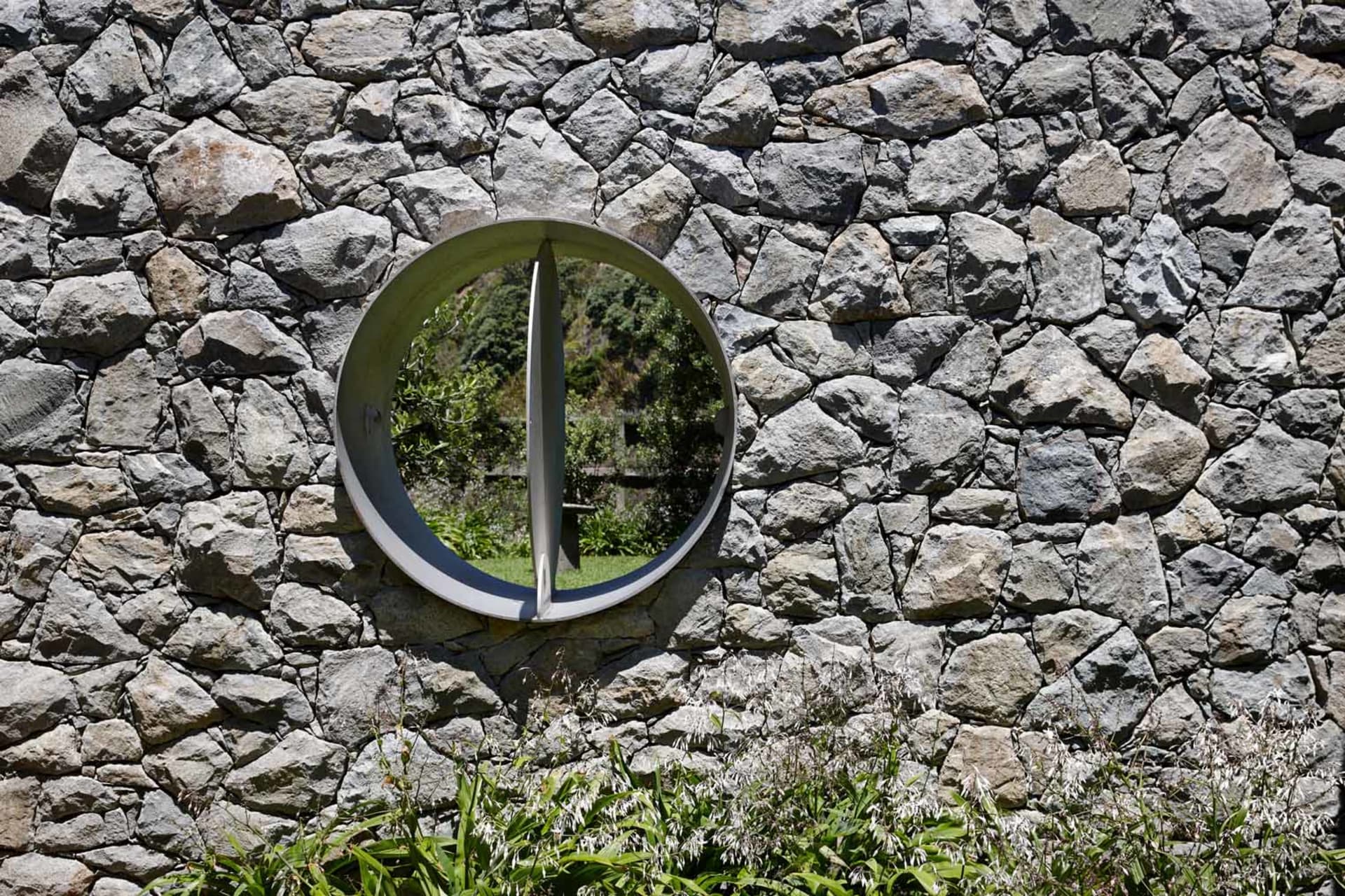 Circular stone wall with a view through it, surrounded by greenery and rocks.