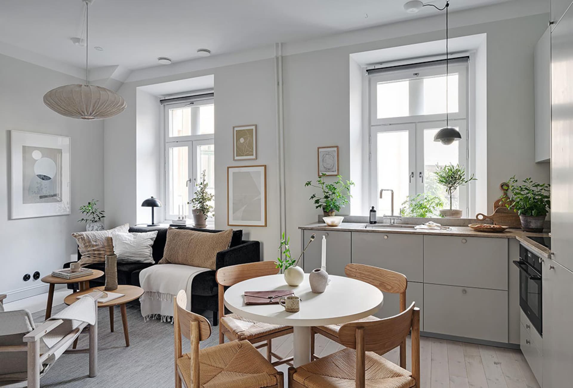 A minimalist residential space featuring a white dining table surrounded by wooden chairs, a grey sofa with white cushions, a