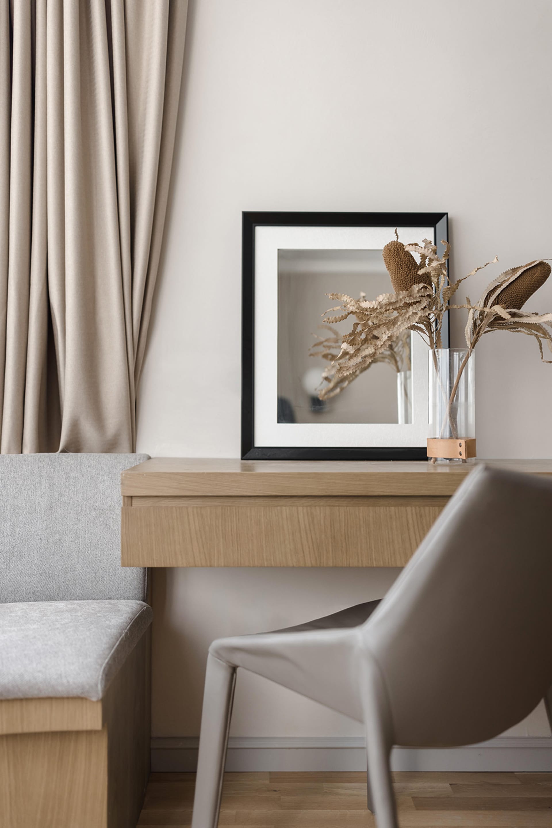 A modern interior workstation featuring a minimalist wooden desk with a black-framed mirror and dried plant decoration.