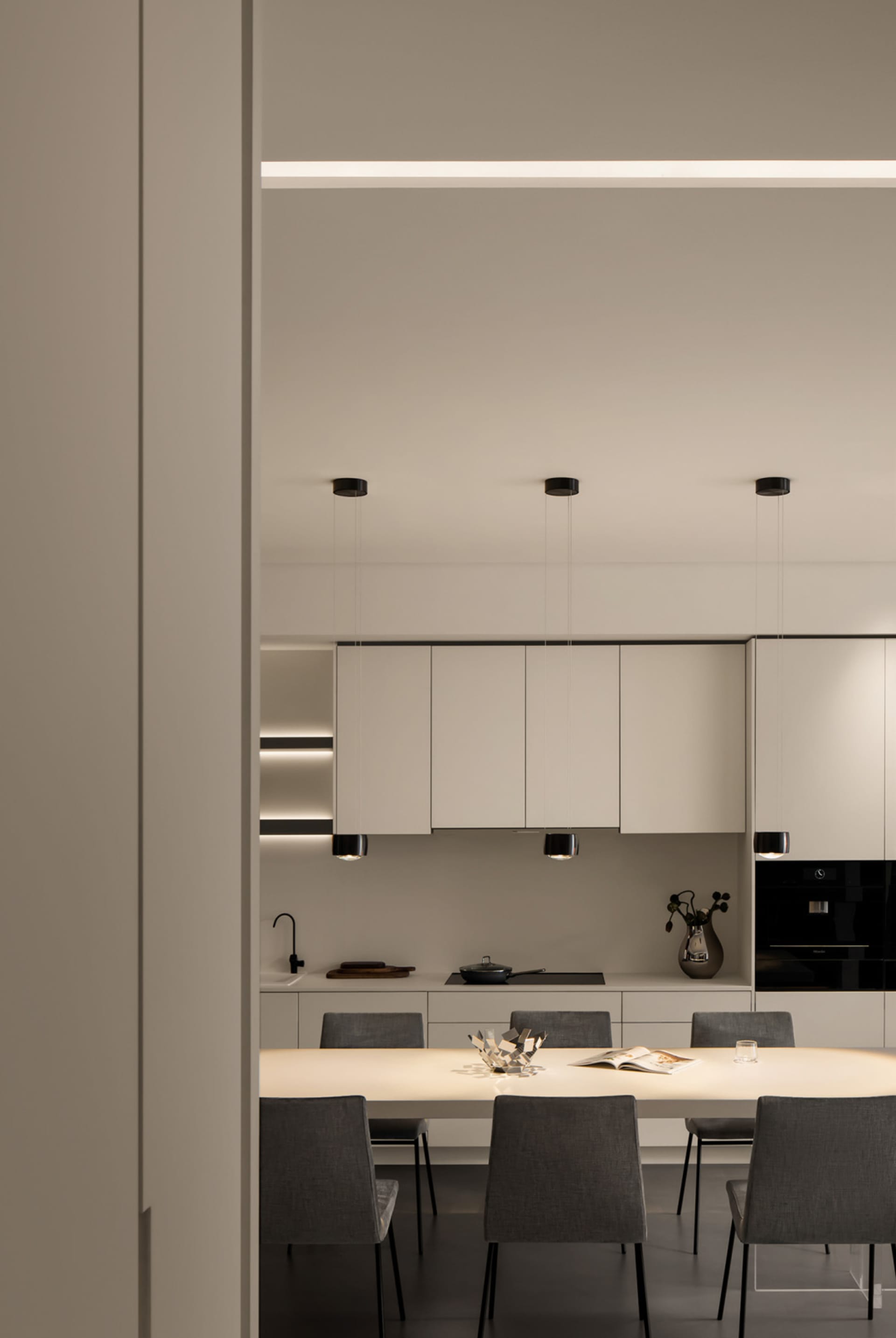 A modern minimalist kitchen with white cabinets, pendant lights, and a dining table.
