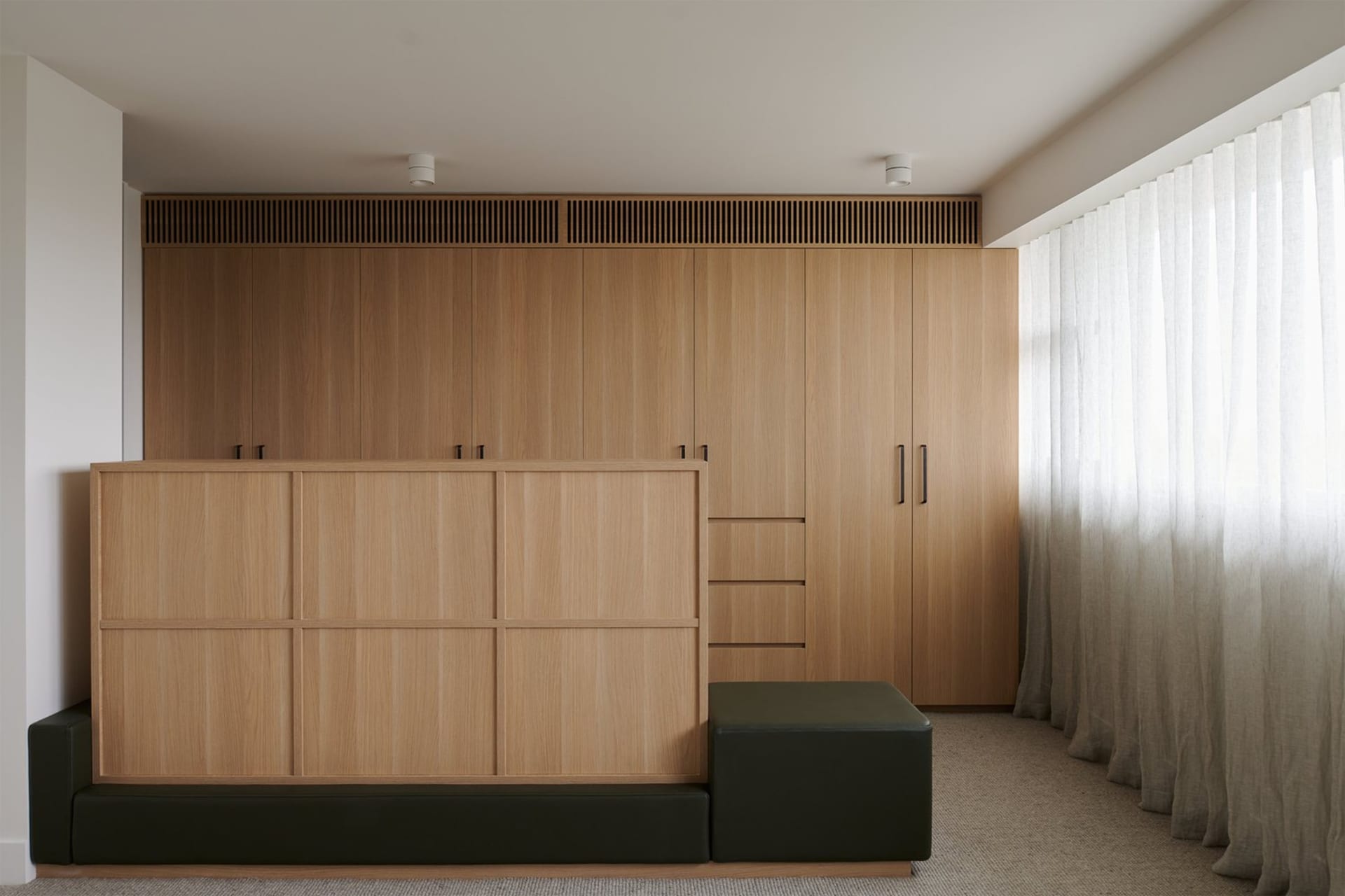 Interior view of a minimalist apartment living room featuring a wooden cabinet wall and a green ottoman.