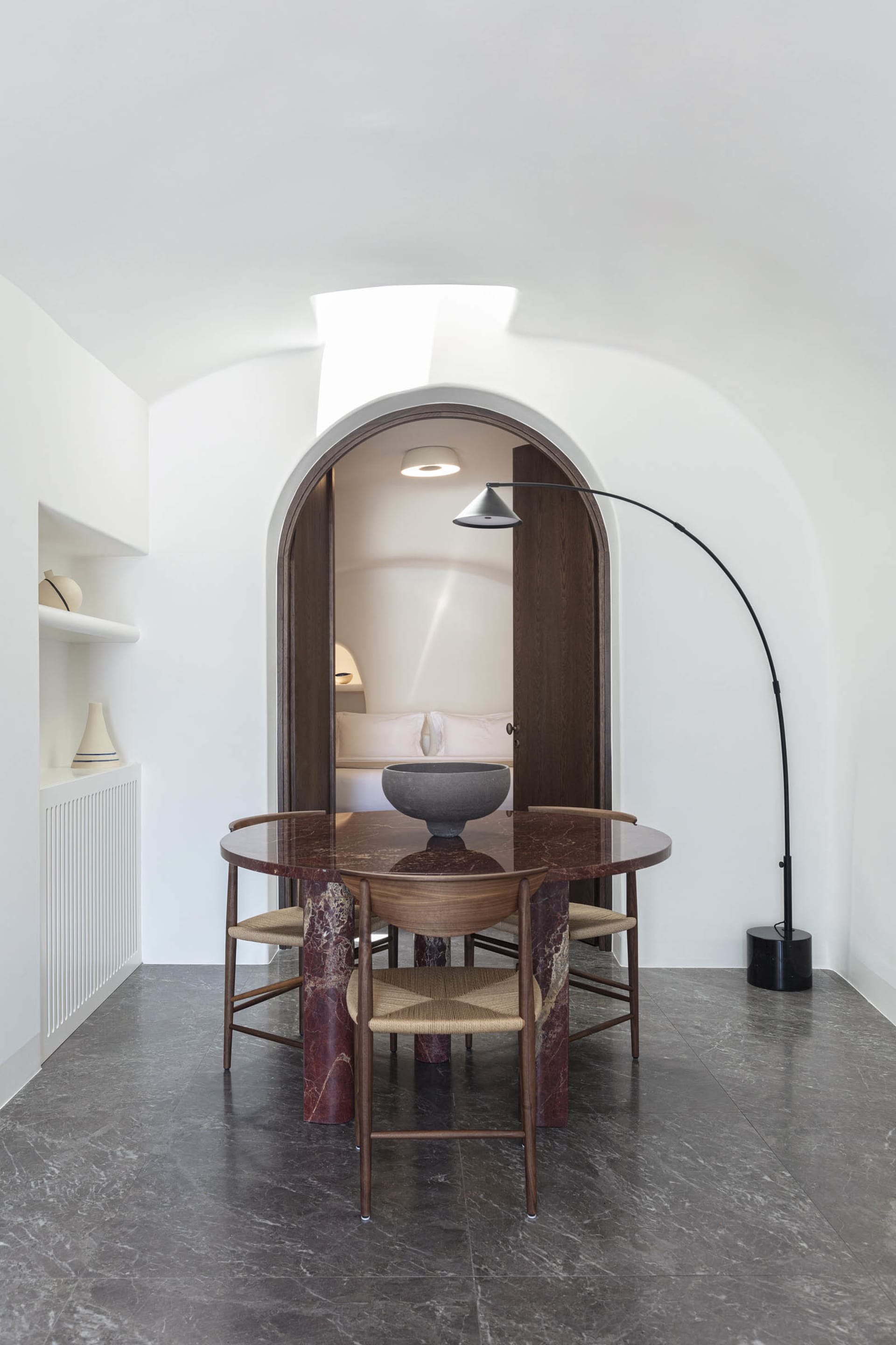 A minimalist dining room with a round table made of red marble and wooden chairs surrounded by white walls and dark grey marb