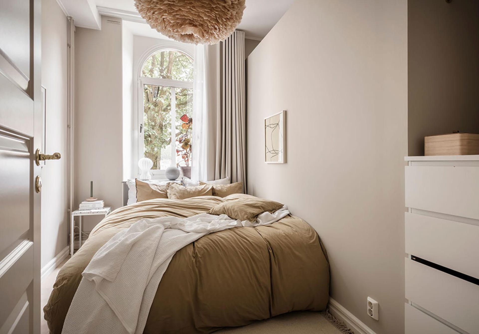 A minimalist bedroom with a bed covered in beige textiles and a floating light fixture above it.
