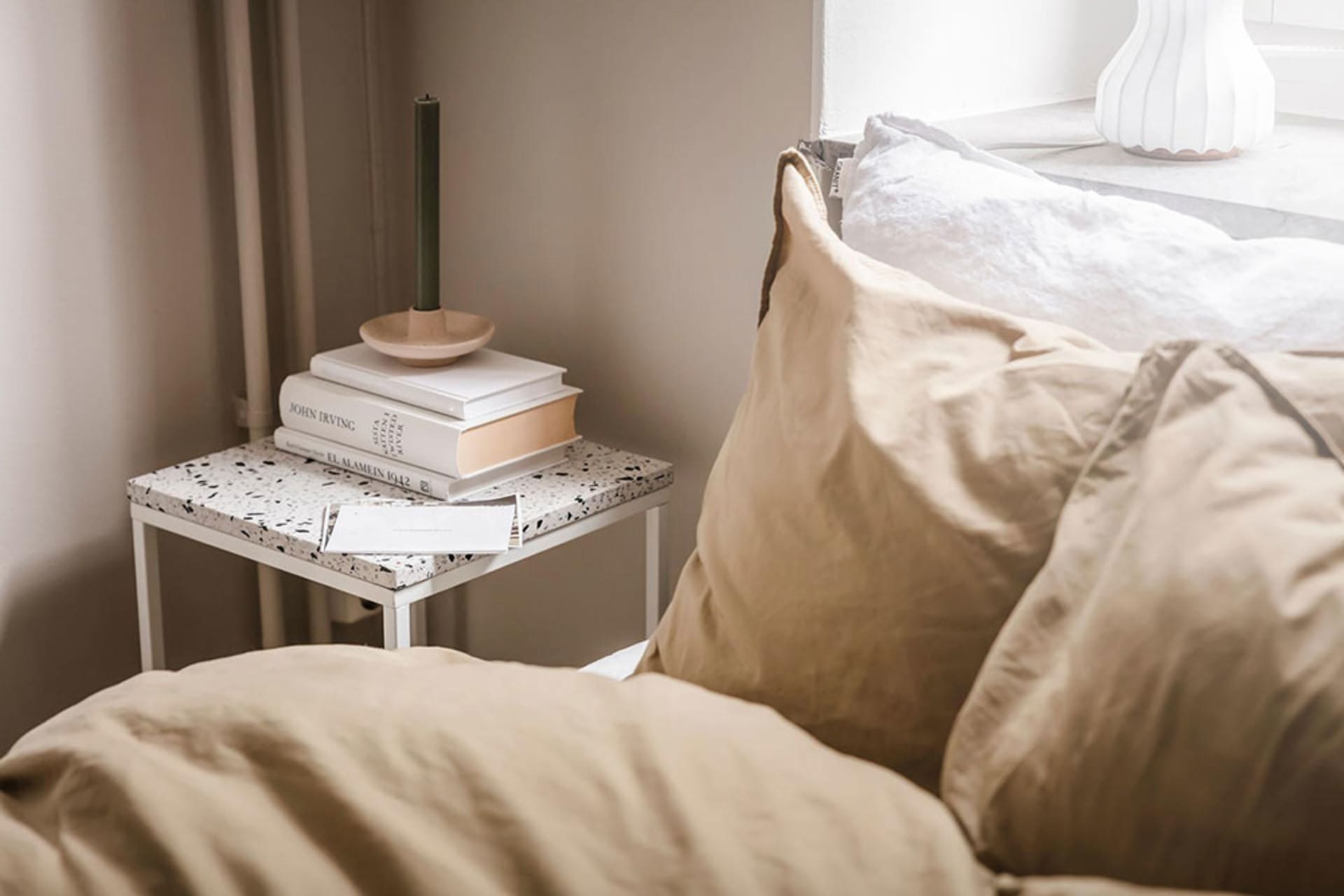 A modern bedroom featuring white textured cotton sheets on a bed, a small side table with books and a candle holder, and a wi