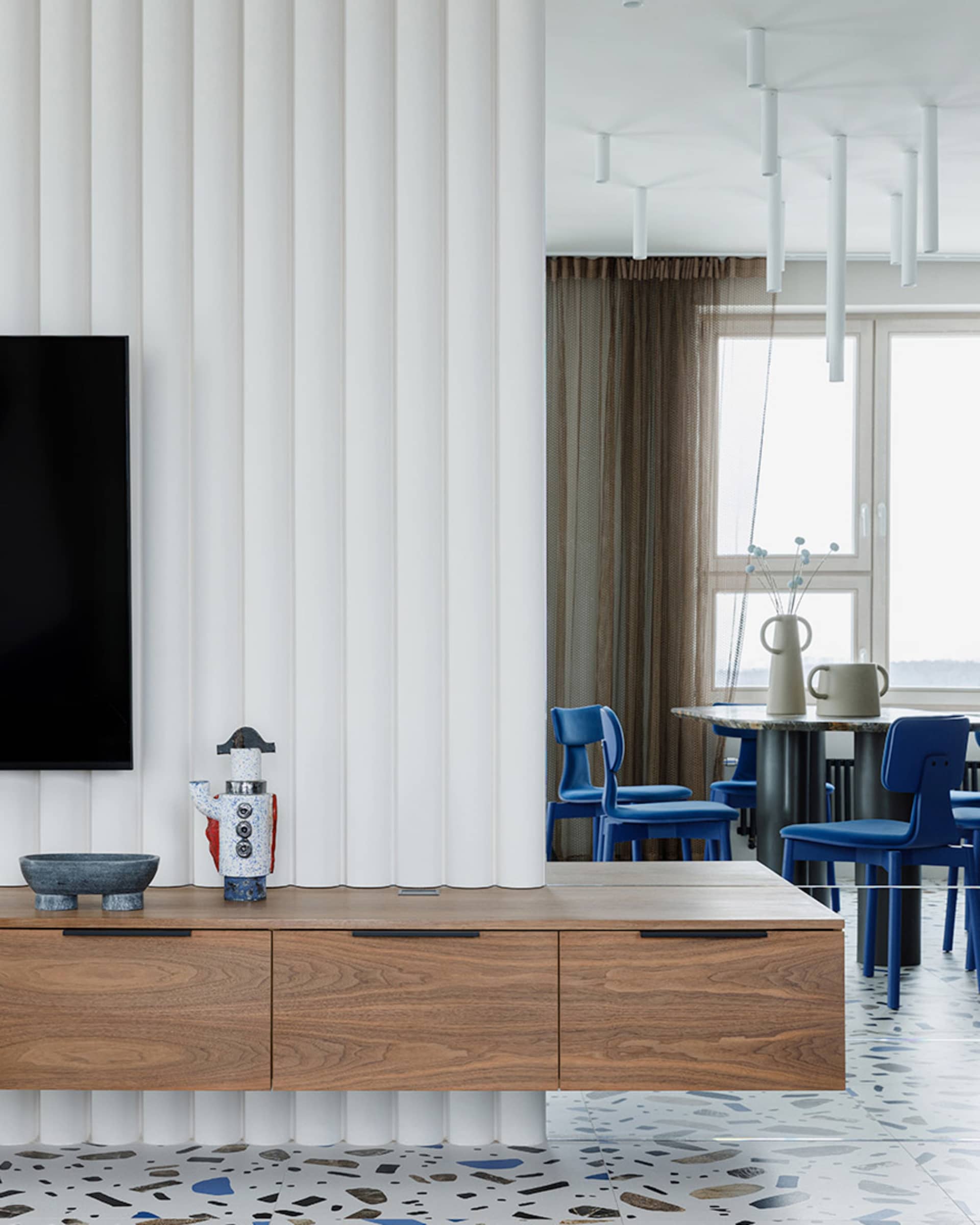 A modern living room with a terrazzo floor, blue chairs, and a wooden floating cabinet.