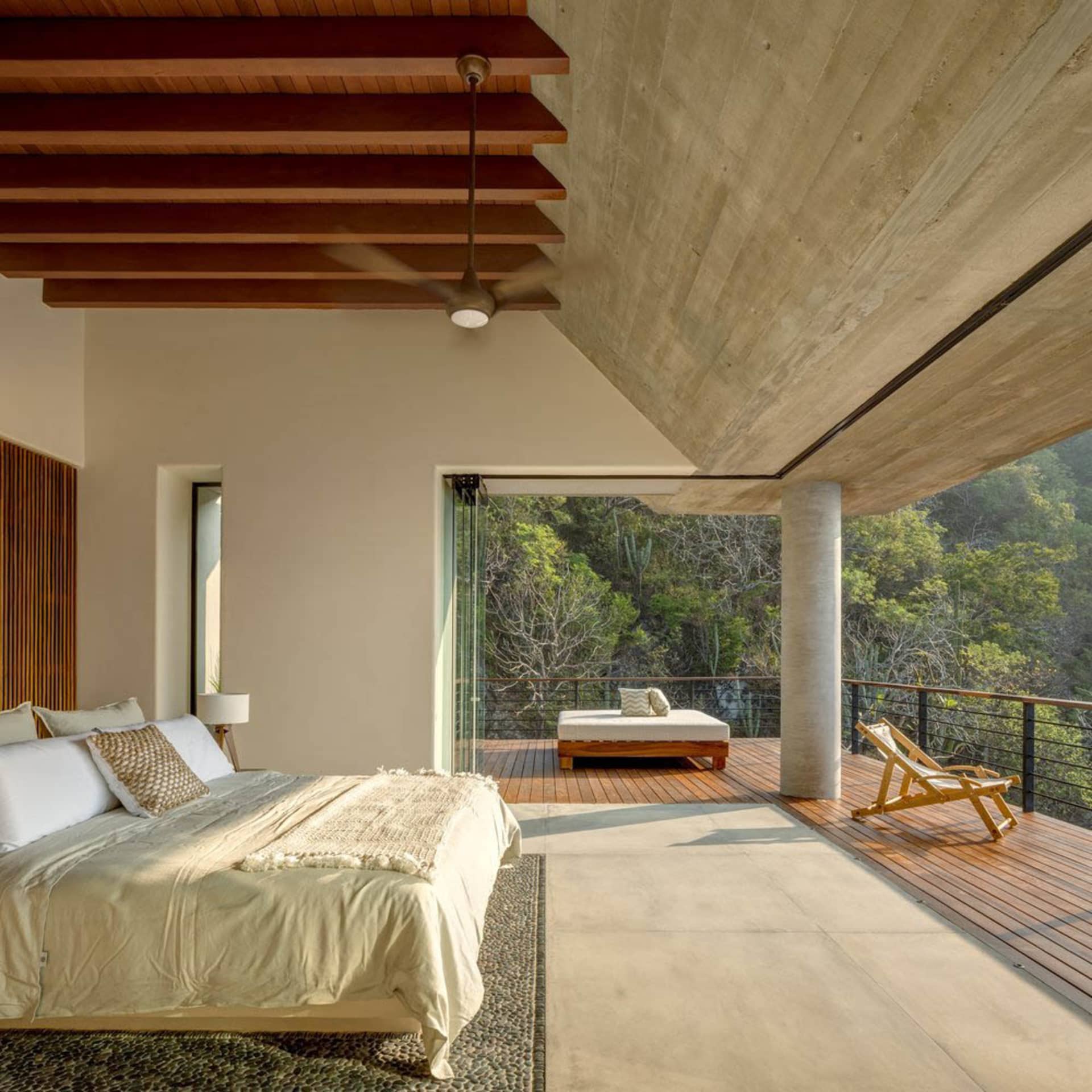 A modern bedroom featuring a concrete ceiling and wooden beams, with a view of lush greenery outside through large windows.