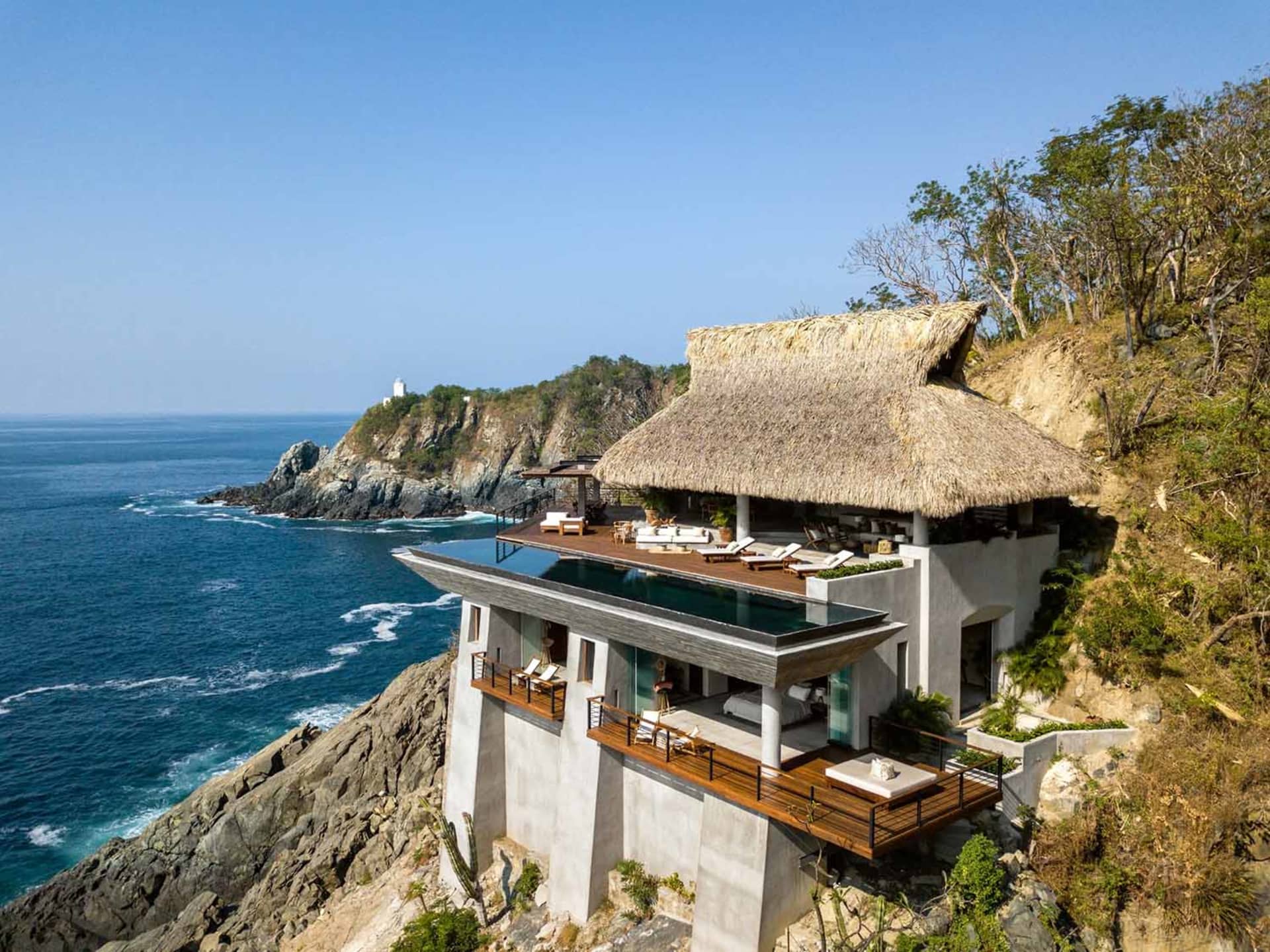 Modern cliffside house with a thatched roof and infinity pool overlooking the ocean.