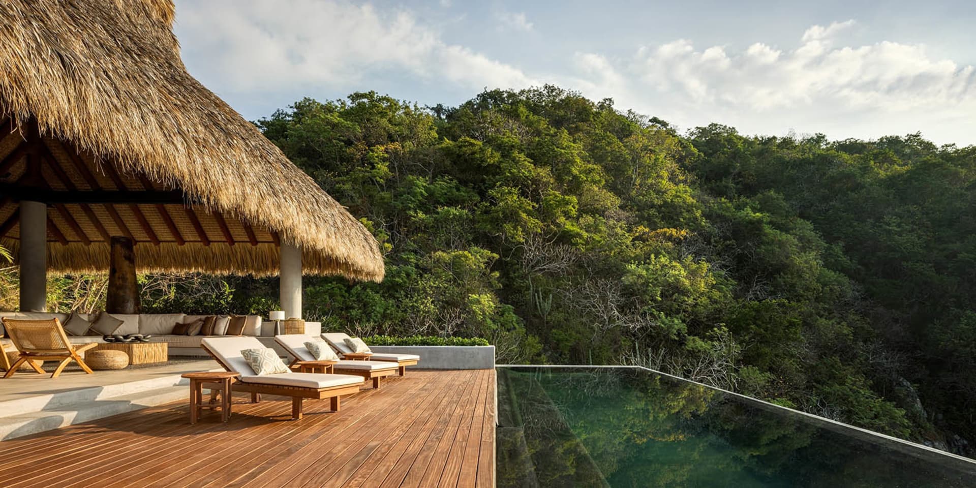 A luxurious outdoor living area with a thatched roof, wooden deck, pool, and lush green surroundings.