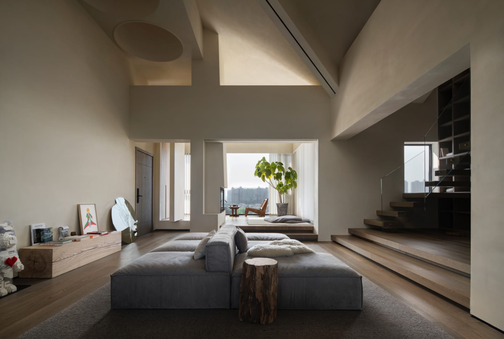 A modern living room with a dark grey modular sofa, a wooden coffee table, and a large window overlooking urban buildings.