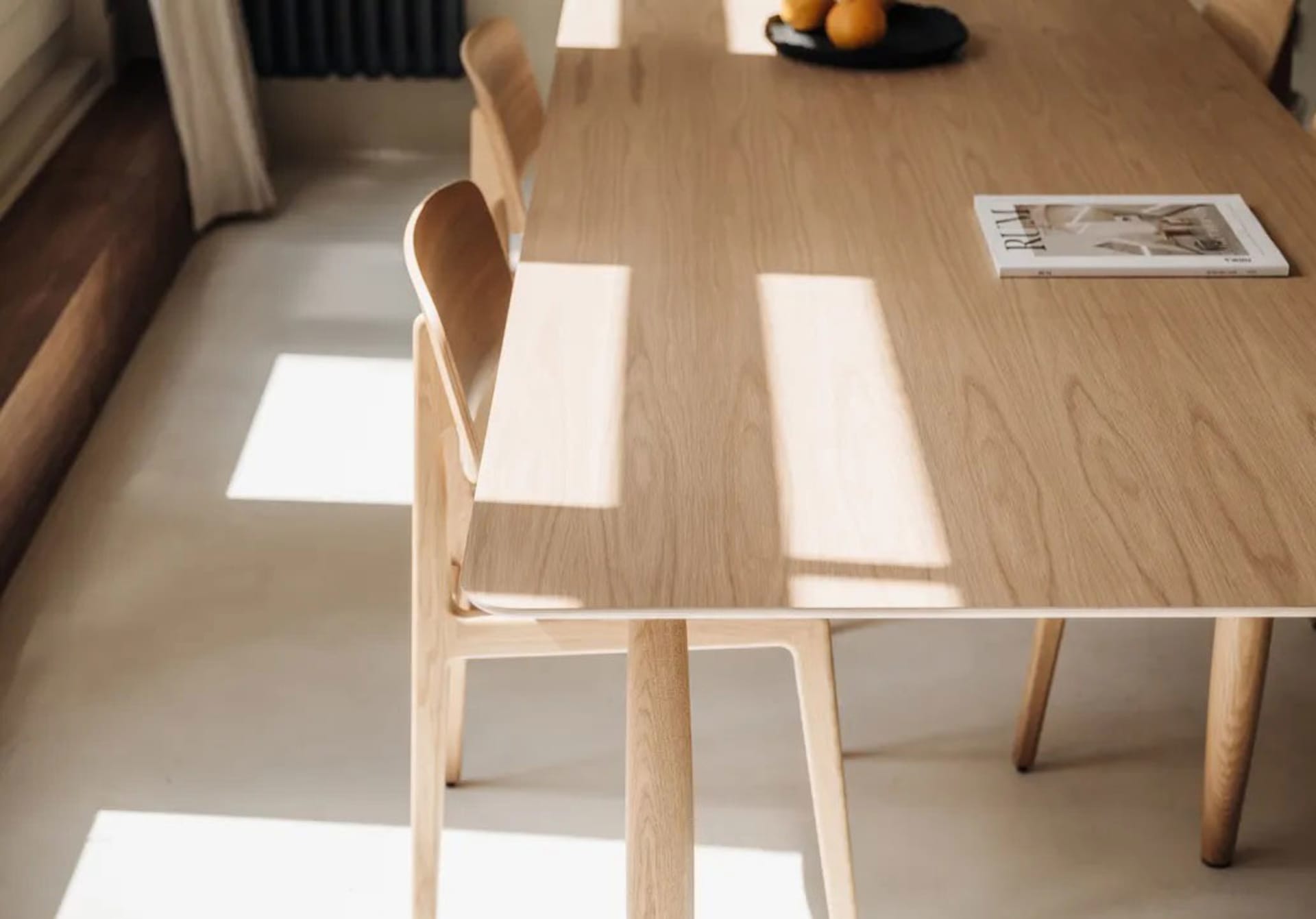A modern interior setting featuring a wooden dining table and chairs, with a magazine and oranges on the table, and sunlight