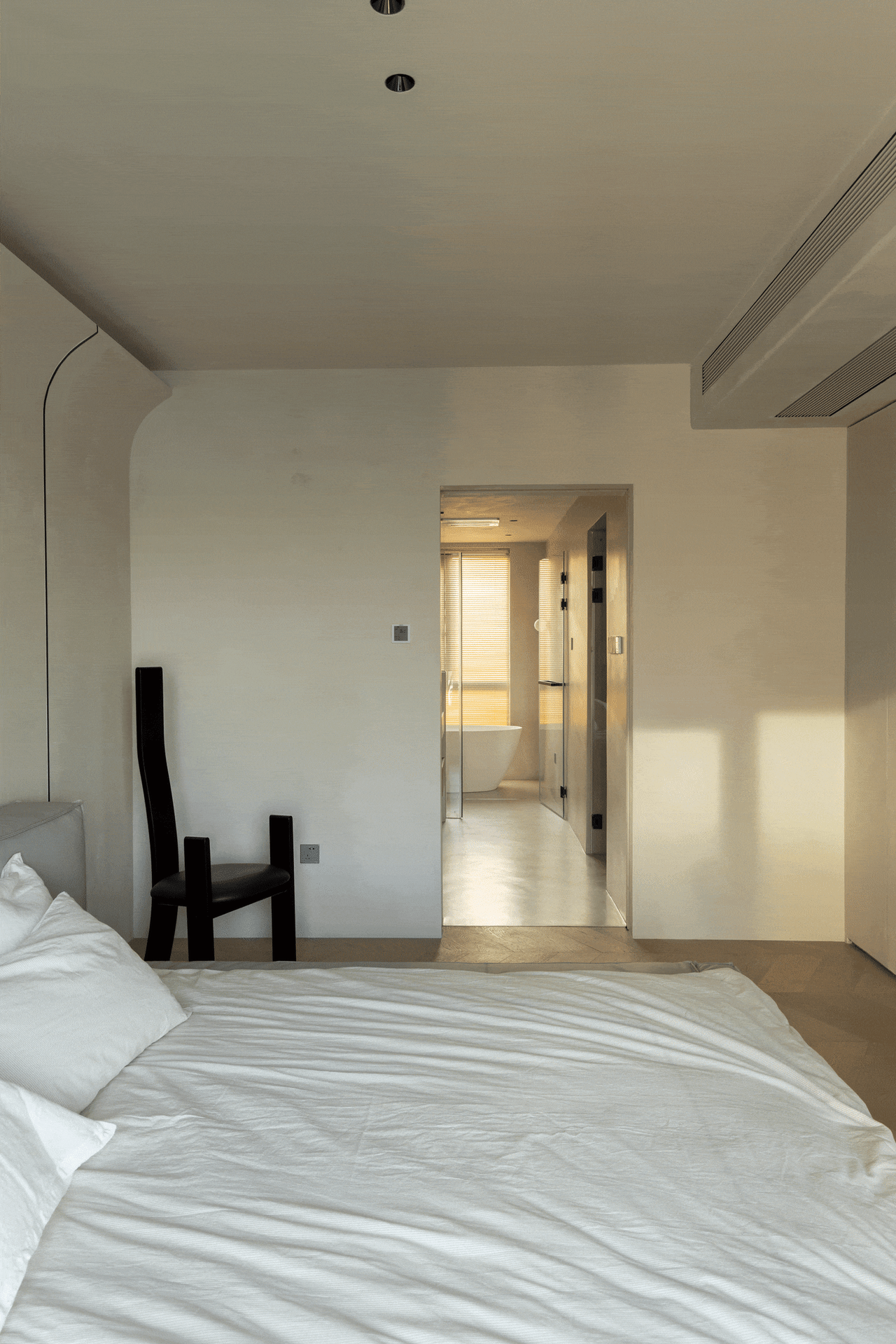 A minimalist bedroom featuring neutral tones with a view into an adjacent bathroom