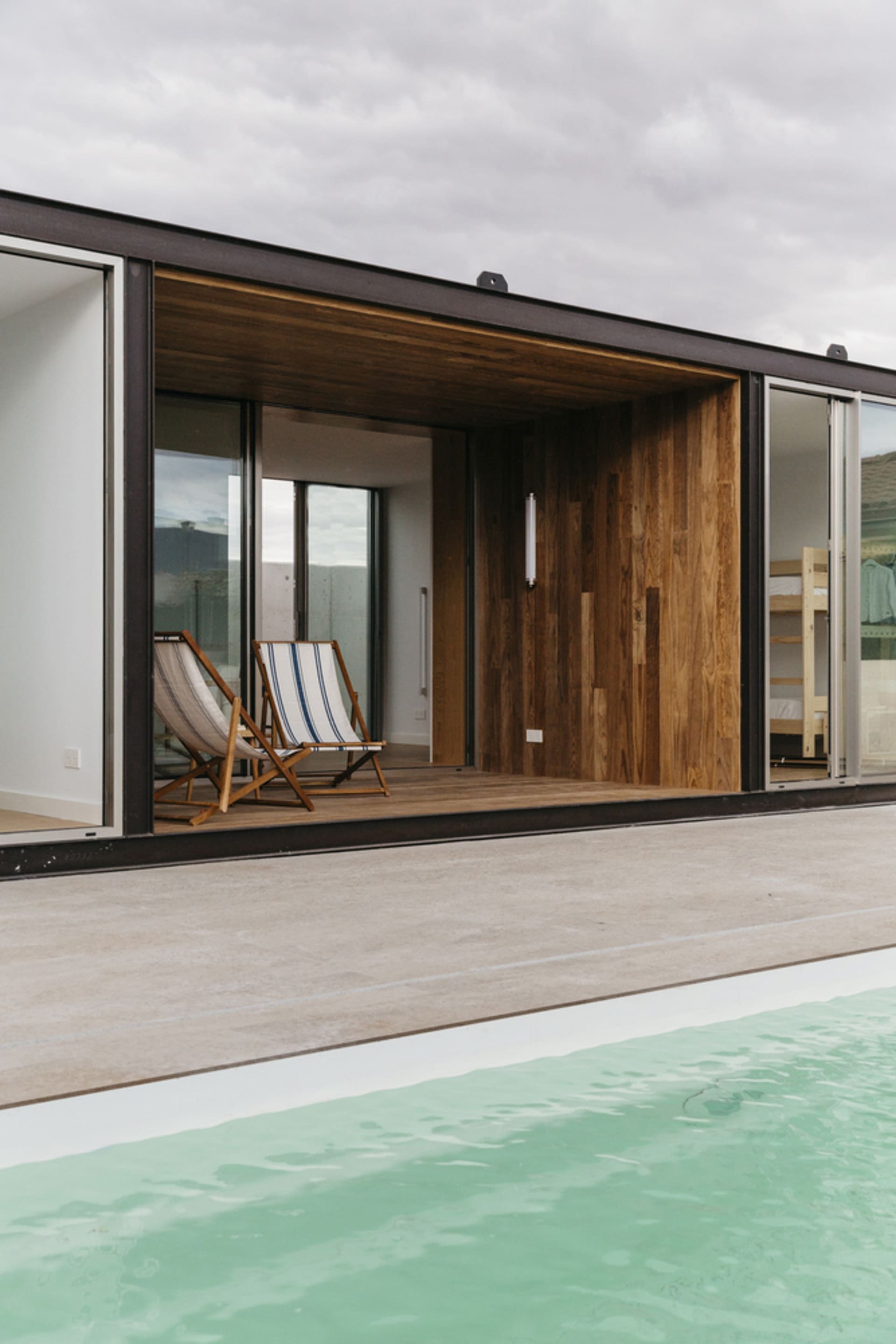 Modern hut with a pool, wooden and glass walls, and two deck chairs.