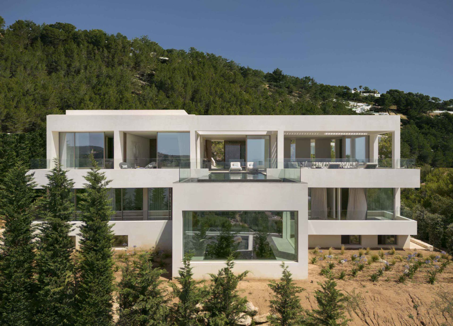 A modern, white residential house with spacious glass walls overlooking a green hillside.