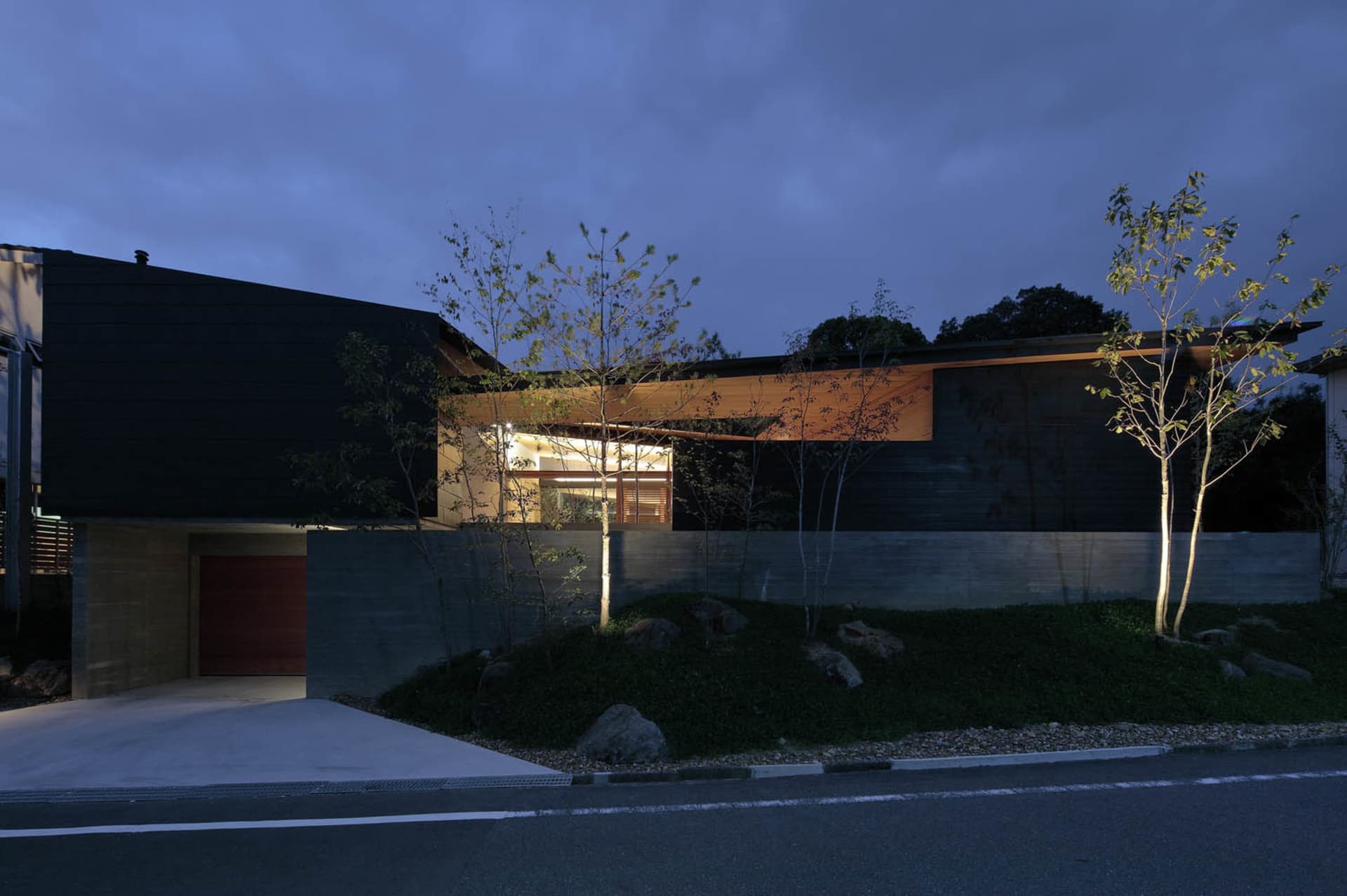 A modern house with a black exterior made of concrete and wood, featuring a driveway and tree lighting.