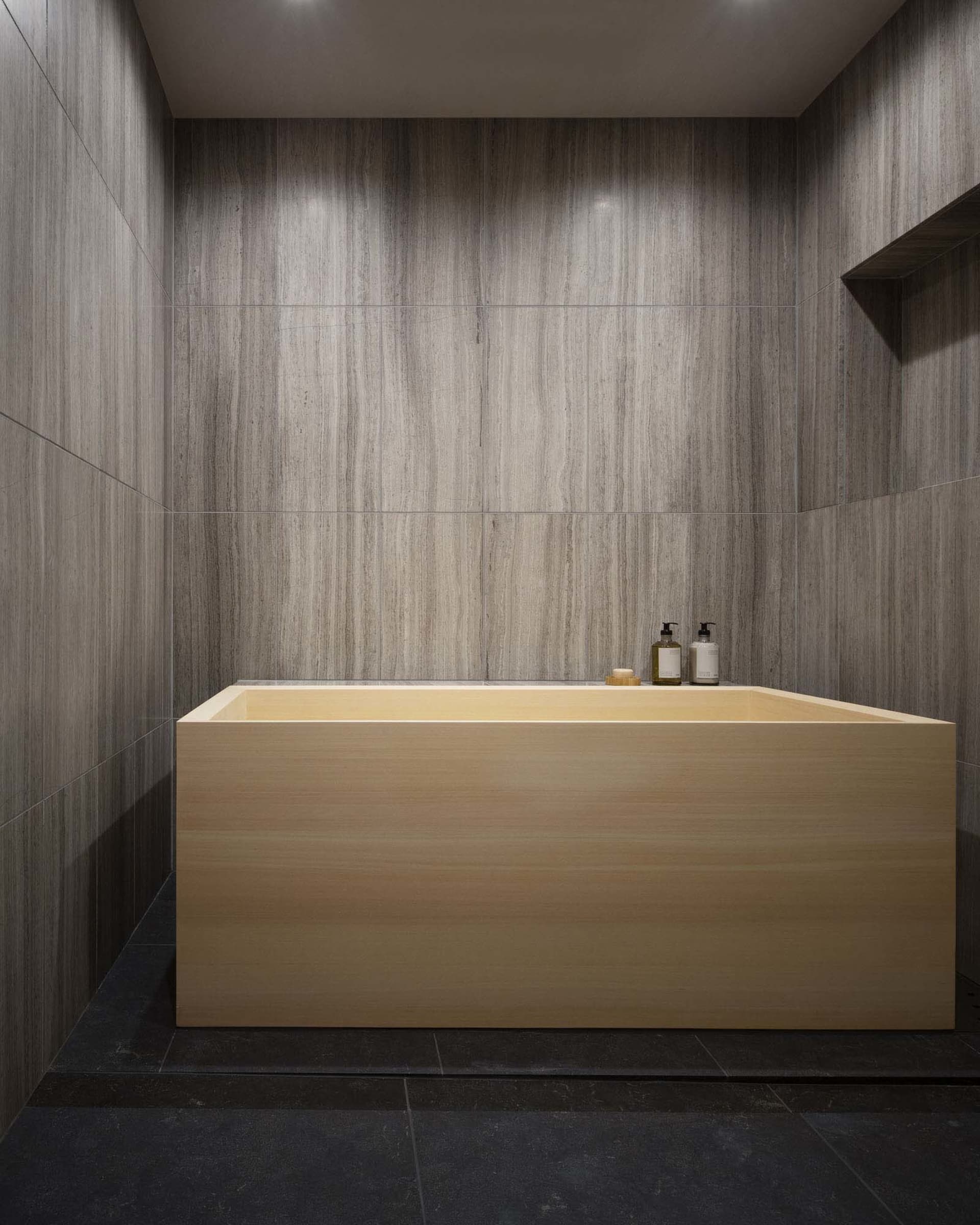 Minimalist modern bathroom with a wooden bathtub, grey stone walls, and black floor tiles.