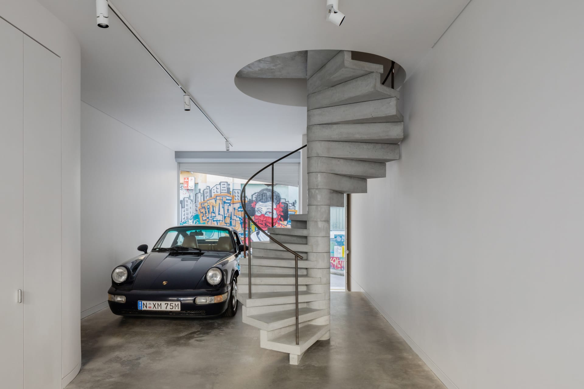 A modern garage with a black Porsche 911 parked next to a concrete spiral staircase.