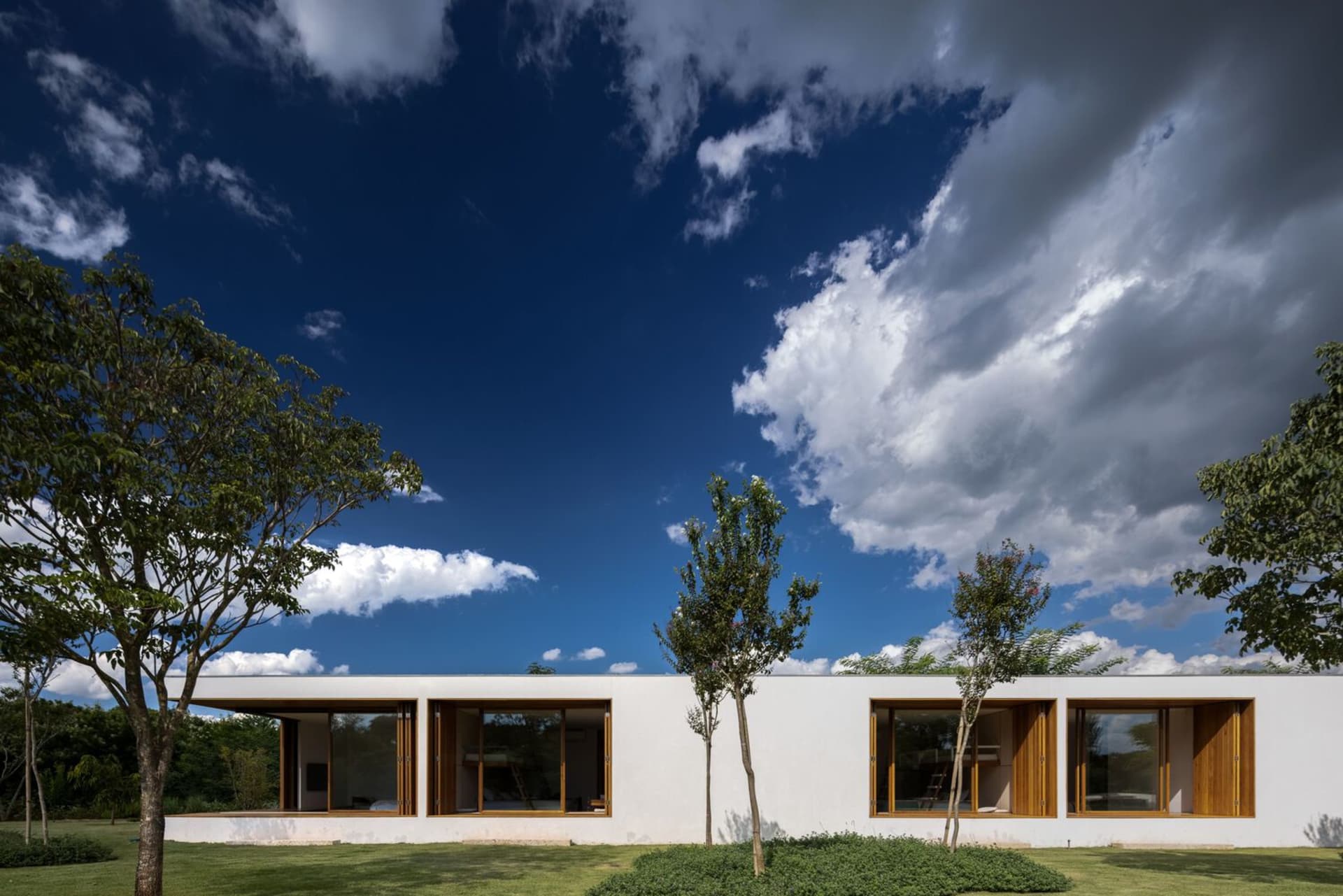 A modern residential building with white plaster exterior, large windows, and a flat roof surrounded by trees and greenery ag