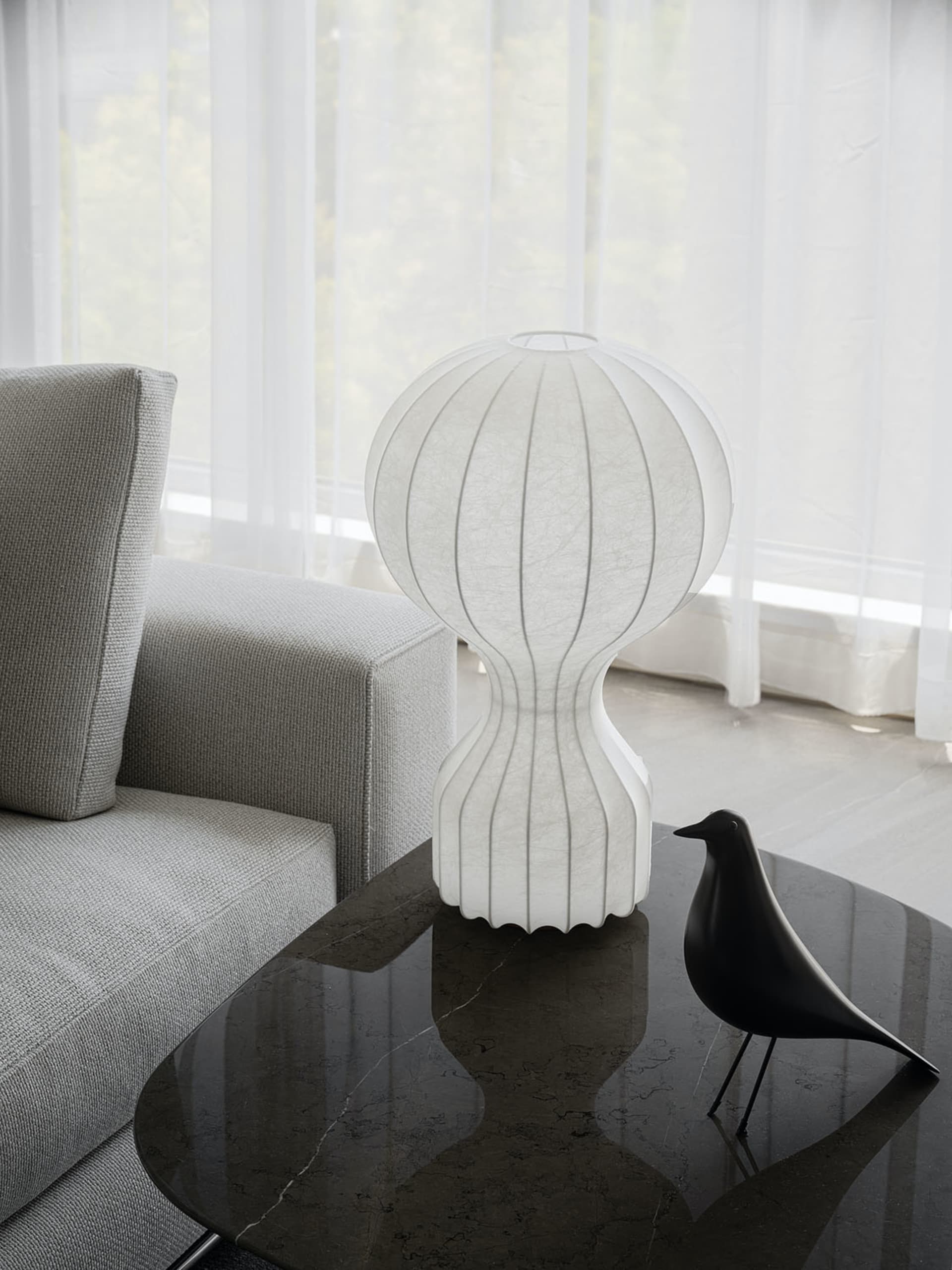 Modern living room with a sleek grey sofa, white fabric lampshade on a glass table, and a black bird figurine.